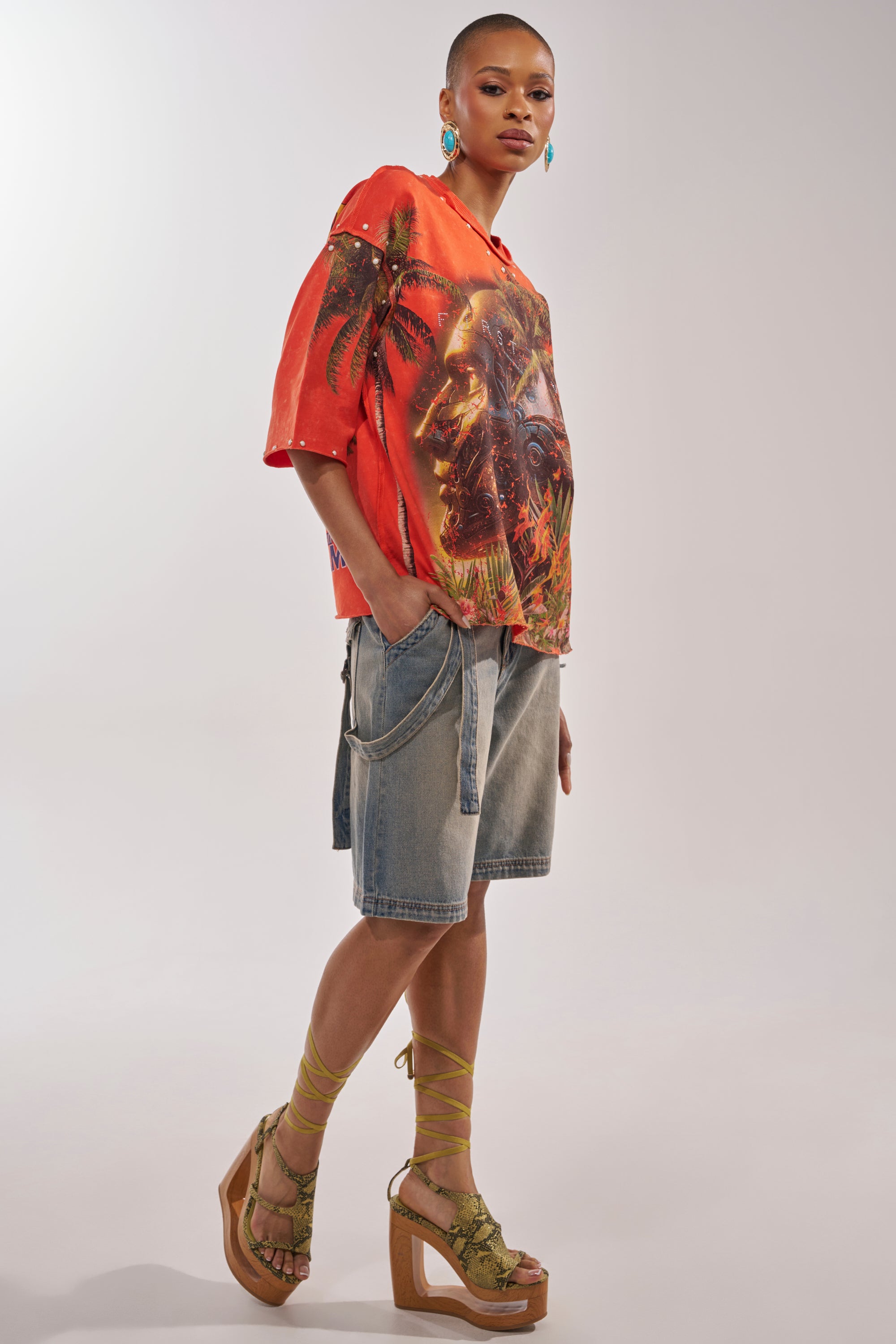 A person in side profile wears the FIRST ROW LOST PARADISE RHINESTONE FRENCH TERRY TEE, paired with loose denim shorts, green lace-up wedge sandals, and turquoise earrings, hands in pockets against a plain background. 5