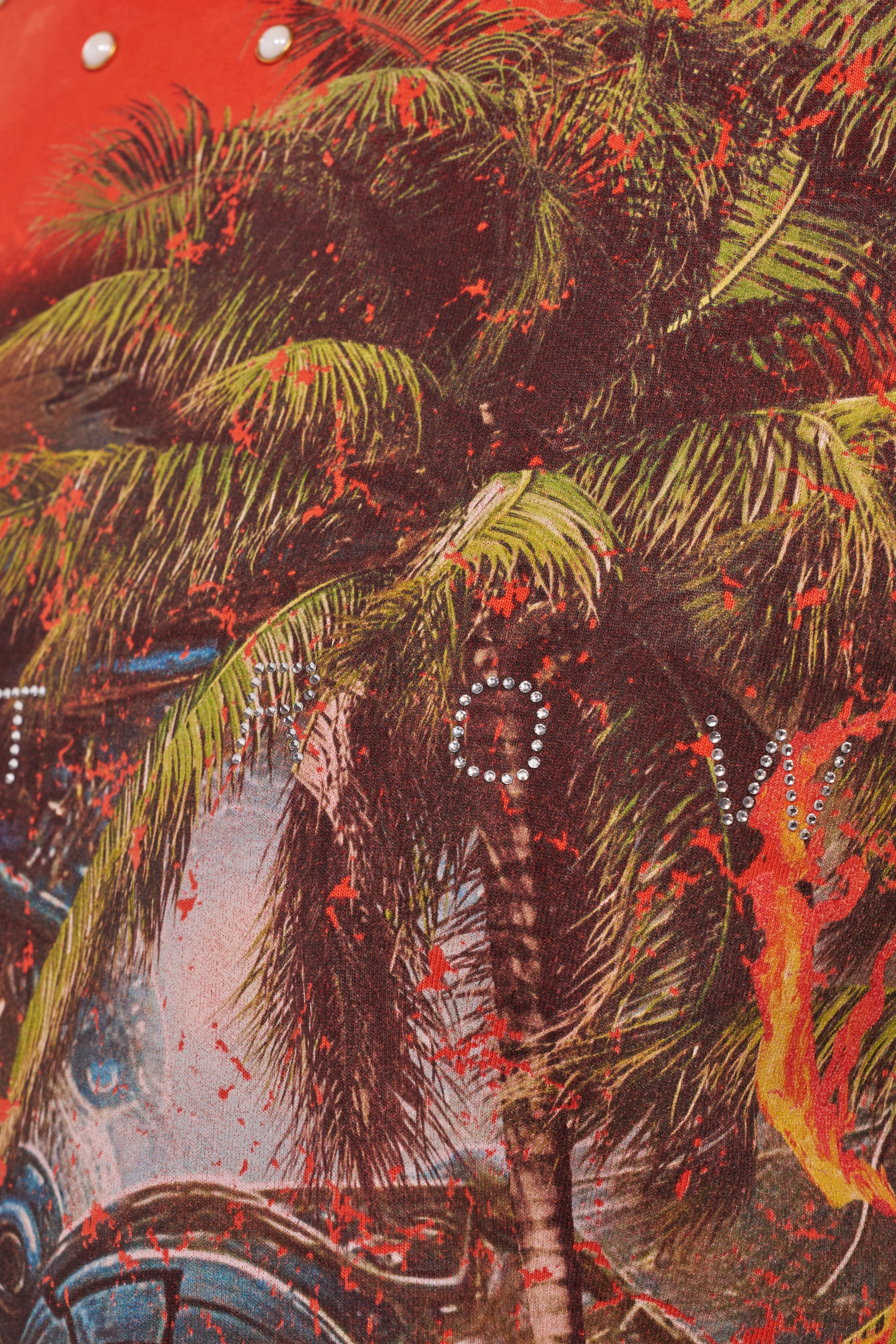 A close-up of the FIRST ROW LOST PARADISE RHINESTONE FRENCH TERRY TEE, showcasing vibrant tropical fabric with palm trees, red splatters, pearl studs, and rhinestone letters on an orange background. 7