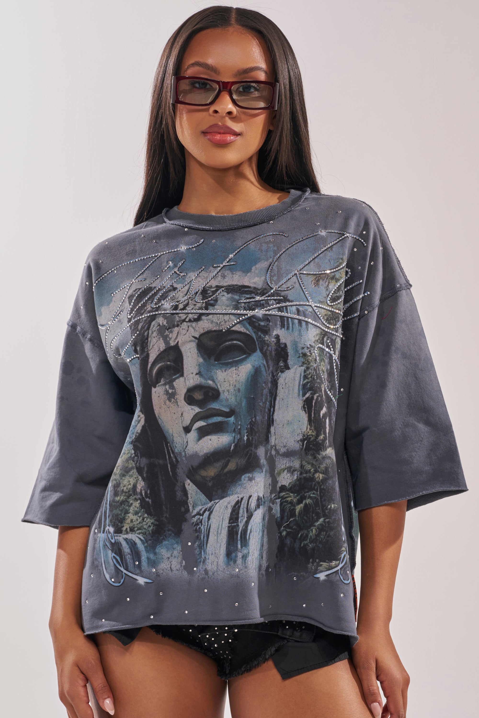 A woman with long straight hair nails street style in sunglasses, black studded shorts, and the FIRST ROW DIVINE RIGHT FRENCH TERRY TEE—a loose dark gray cropped top with a graphic stone face and waterfalls. 0