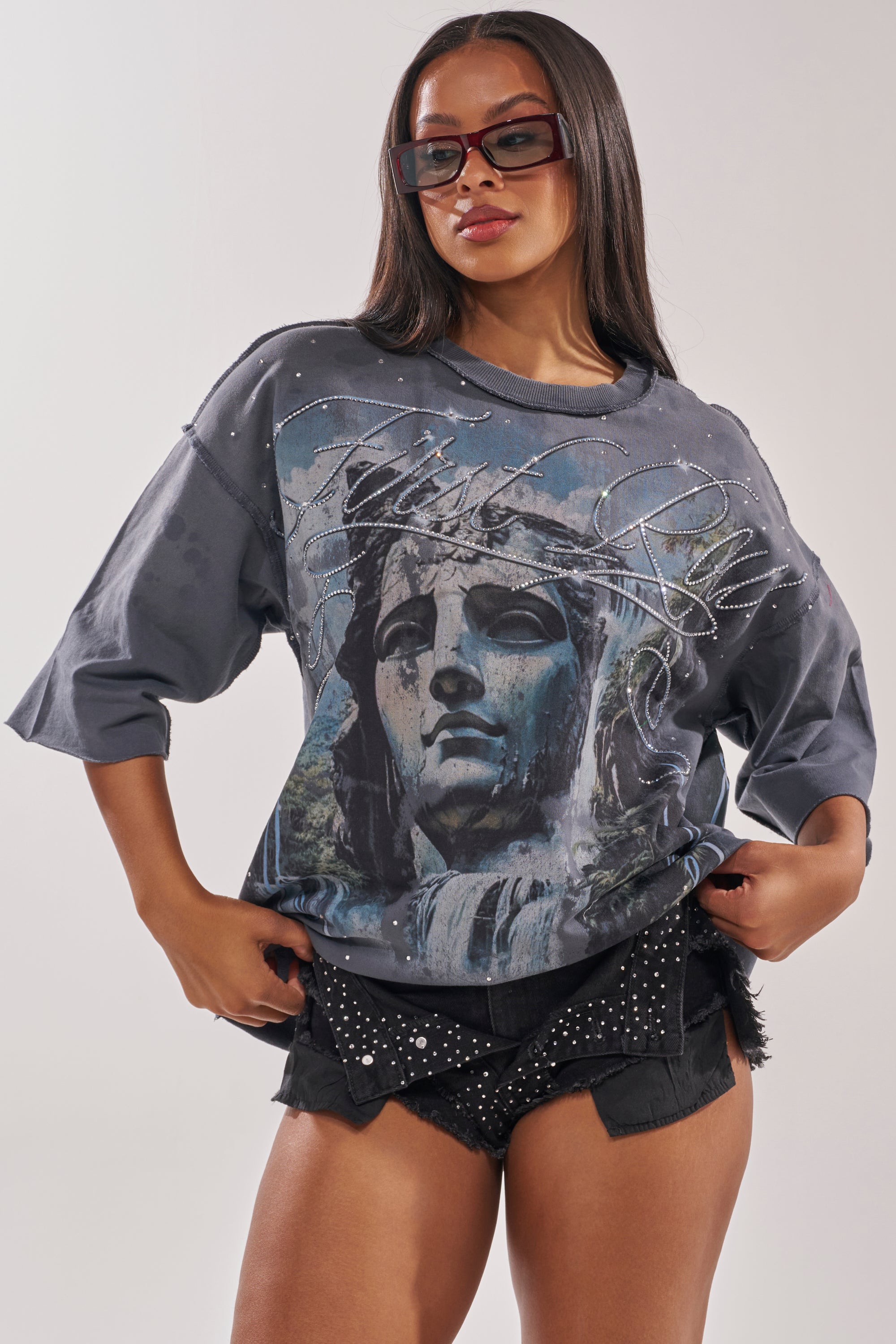 A woman showcases street style in oversized sunglasses, the FIRST ROW DIVINE RIGHT FRENCH TERRY TEE with a statue graphic and rhinestones, and black studded shorts as she poses with her hands in her pockets. 1