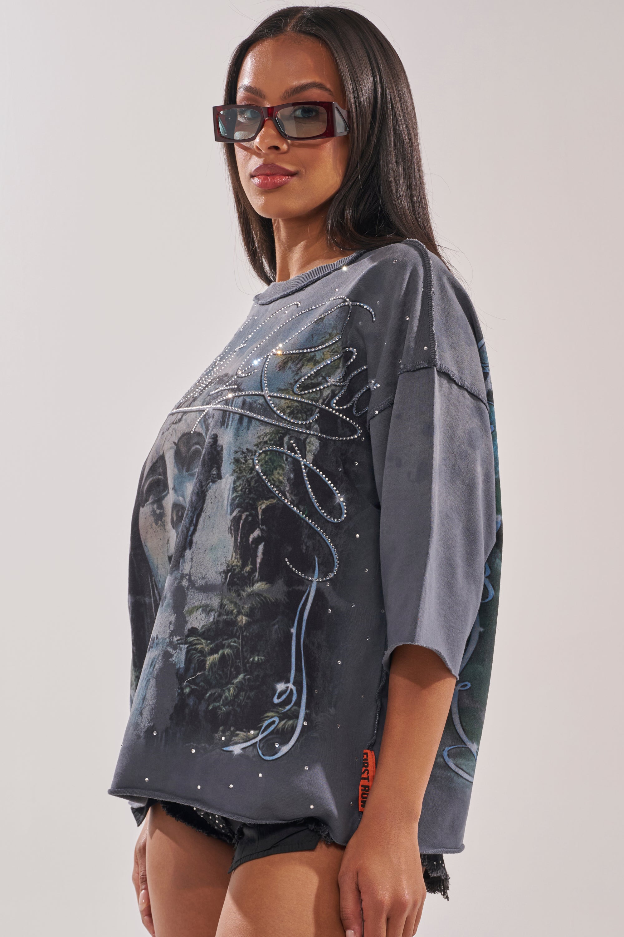 A woman with long straight hair and sunglasses channels street style in the FIRST ROW DIVINE RIGHT FRENCH TERRY TEE, an oversized graphic cropped top with rhinestones, paired with black shorts and posed against a plain background. 2