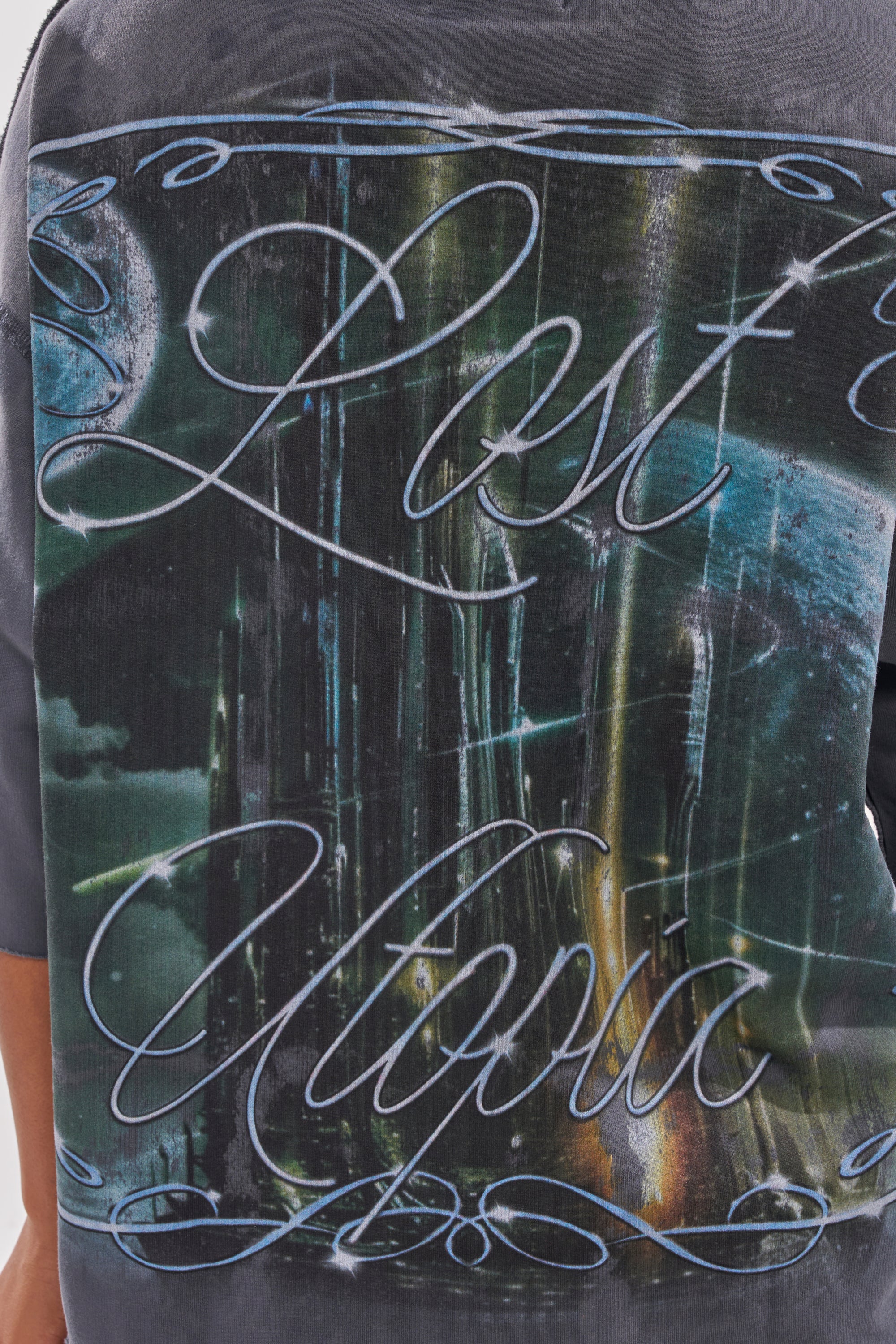A close-up of the FIRST ROW DIVINE RIGHT FRENCH TERRY TEE featuring stylized "Lost Utopia" cursive text with rhinestone accents over a dark blue and green abstract background. 5