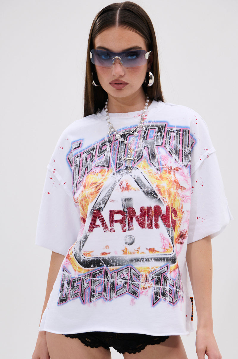 A woman models the WARNING SIGNS CROPPED GRAPHIC TEE, a loose white crop top with bold, distressed "WARNING" graphics on the front, styled with blue sunglasses, silver earrings, a chunky chain necklace, and black shorts.