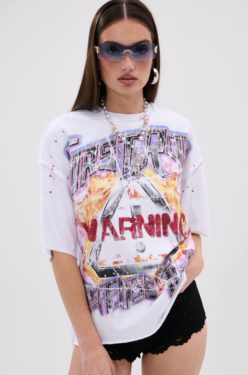 A person with long brown hair wears silver sunglasses, a chunky silver necklace, black lace shorts, and the WARNING SIGNS CROPPED GRAPHIC TEE with flame distressed graphics and "WARNING" signs, posing against a plain background.