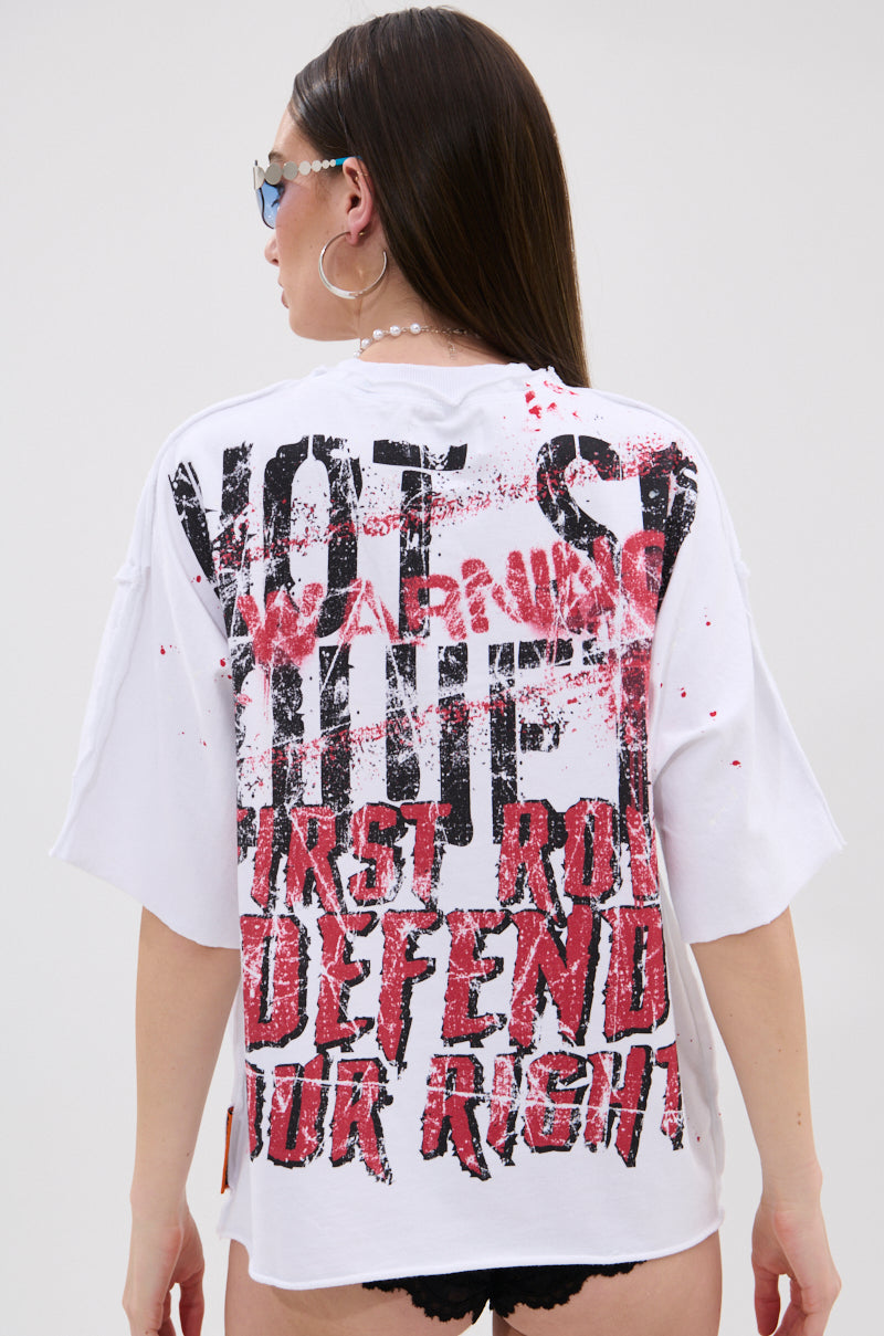 A woman with long brown hair and sunglasses is seen from behind, wearing the WARNING SIGNS CROPPED GRAPHIC TEE, a white oversized shirt with bold distressed black and red graphics reading "NOT WARNING FIRST, ROLL DEFEND YOUR RIGHT.