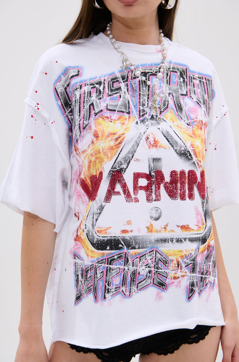 A woman wears the WARNING SIGNS CROPPED GRAPHIC TEE, a white oversized shirt with distressed warning sign graphics and splatter designs. She completes her edgy look with a silver chain necklace and black shorts.