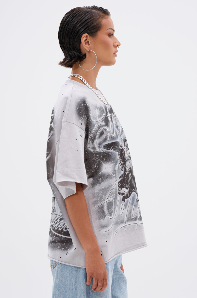 A person with short, slicked-back hair, large hoop earrings, a chunky chain necklace, light blue jeans, and the BOUNCE BACK OVERSIZED TEE stands in profile against a plain white background.
