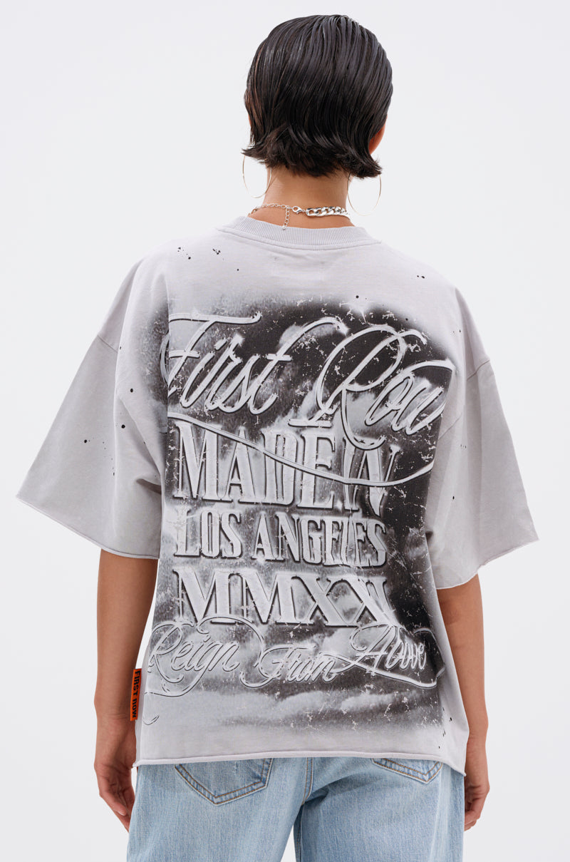 A person with short dark hair, wearing a silver chain, stands facing away. They wear the BOUNCE BACK OVERSIZED TEE with bold graffiti-style text—“First Class, MADE4, Los Angeles, MMXX, Reign From Above”—and light blue jeans.