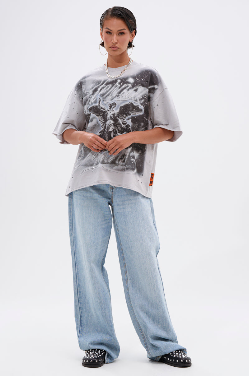 A person stands facing forward against a white background, wearing the BOUNCE BACK OVERSIZED TEE, light blue baggy jeans, black studded shoes, large hoop earrings, and a necklace.