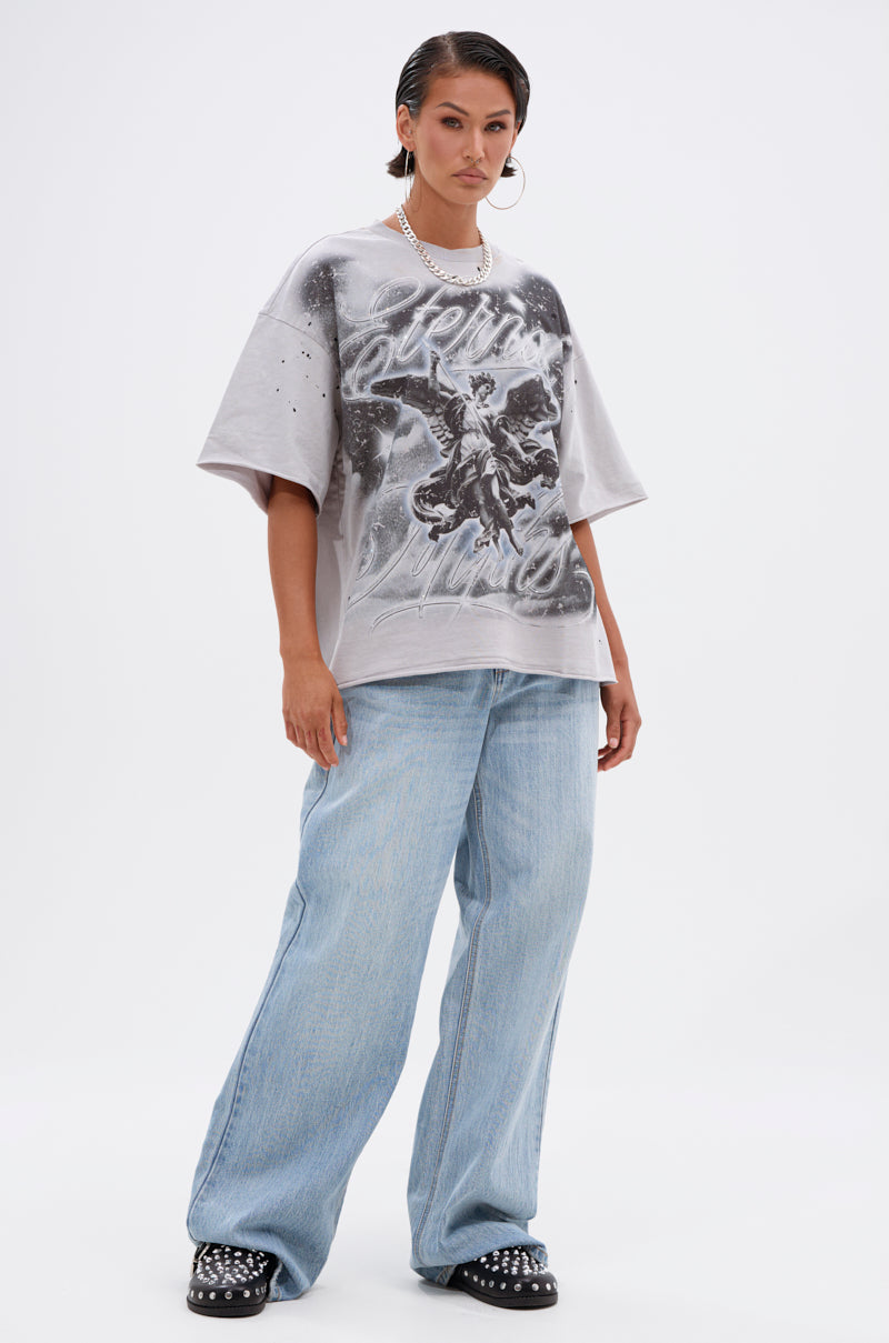 A woman stands confidently against a plain white background wearing the BOUNCE BACK OVERSIZED TEE with black and white graphic art, paired with baggy light blue jeans, black studded shoes, hoop earrings, and a beaded necklace.