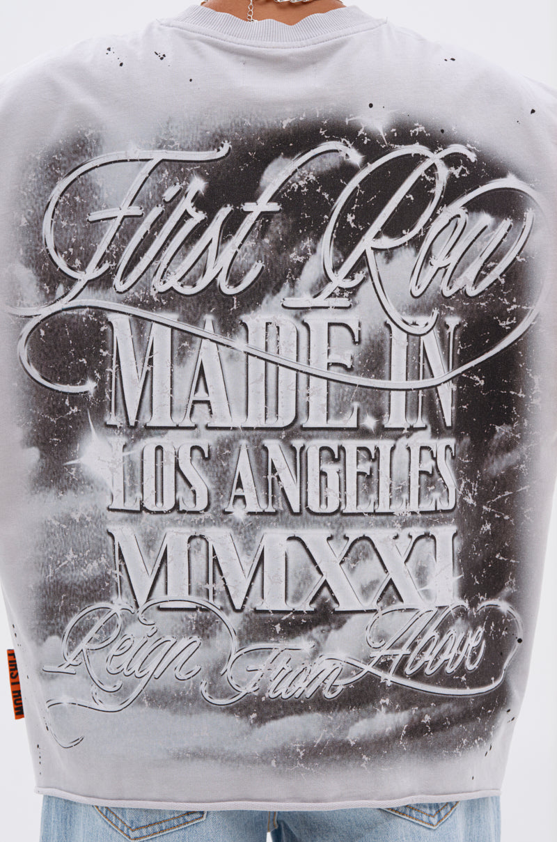 A close-up of the gray BOUNCE BACK OVERSIZED TEE, featuring bold white graffiti-style text: “First Row MADE IN LOS ANGELES MMXXI Reign From Above.” The wearer faces away, showing only the graphic on their back.