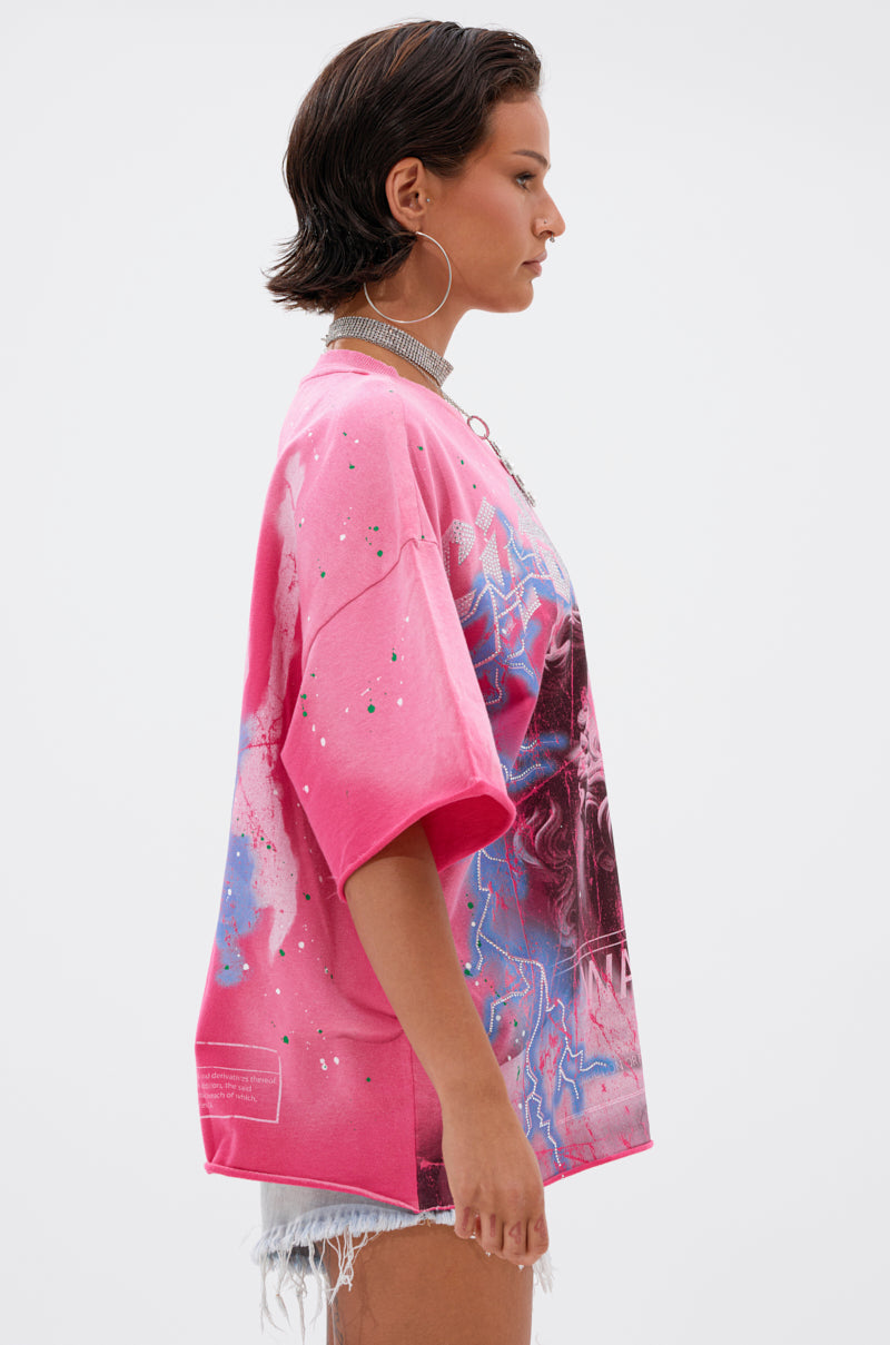 A person with short dark hair, large hoop earrings, a sparkly choker, the FADE INTO BLACK OVERSIZED TEE in bright pink rhinestones, and frayed light denim shorts stands in profile against a plain light background.