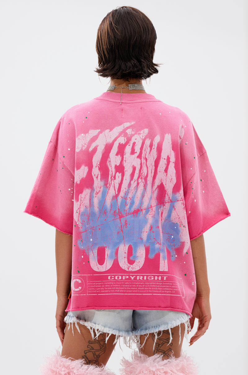 Person with short brown hair wears the FADE INTO BLACK OVERSIZED TEE in bright pink, featuring bold white and blue graffiti-style "ETERNAL" and "COPYRIGHT" text on the back, styled with frayed denim shorts and pink accessories for a standout streetwear look.