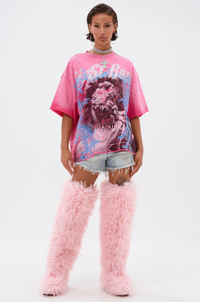 A woman stands facing forward in the FADE INTO BLACK OVERSIZED TEE, styled with frayed denim shorts, chunky necklaces, and tall fluffy pink boots. Her wet-look, slicked-back hair and confident expression stand out against a plain white background.
