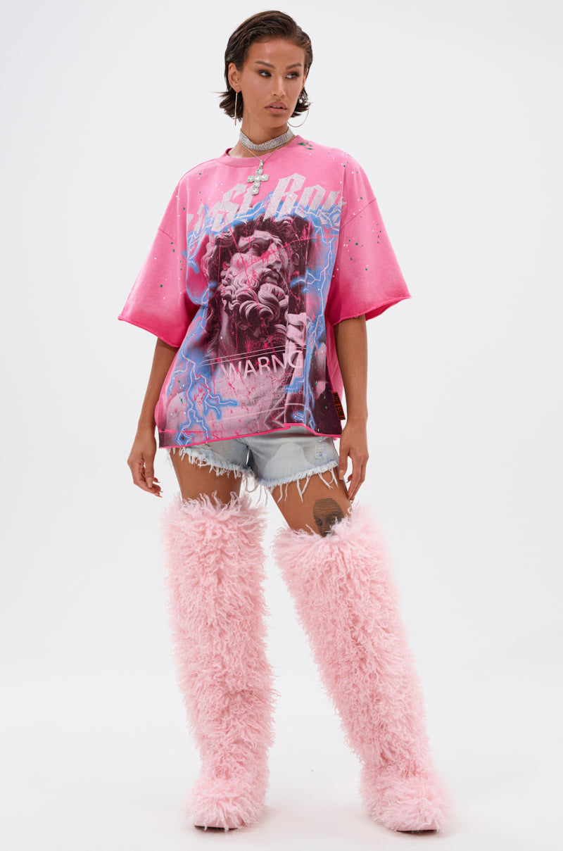 A person with short dark hair and a confident look stands against a white background, wearing the FADE INTO BLACK OVERSIZED TEE in pink, paired with distressed denim shorts and tall, fluffy pink boots.