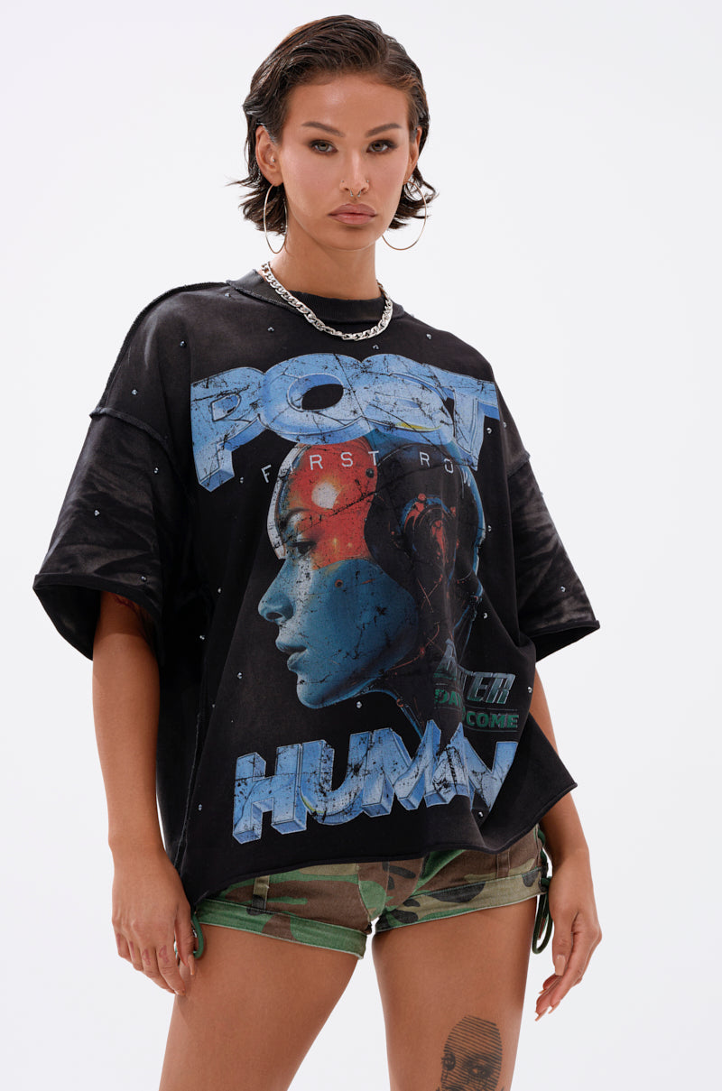 A person with short, wet-look hair wears THE FUTURES CALLING FRENCH TERRY GRAPHIC TEE, a black oversized shirt with a futuristic blue face graphic and "POST HUMAN" text, paired with camouflage shorts and a silver chain necklace.