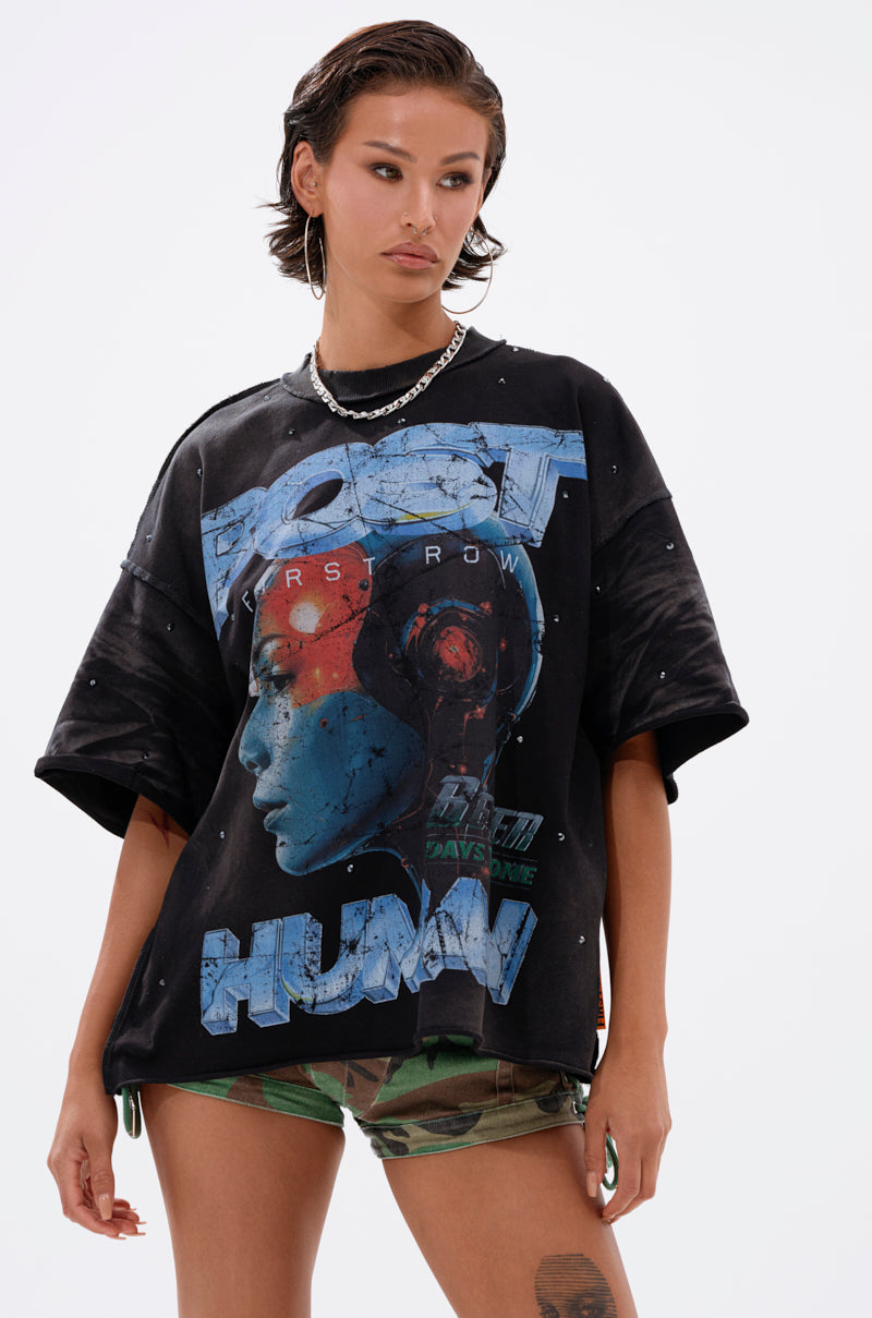 A woman with short brown hair and hoop earrings wears THE FUTURES CALLING FRENCH TERRY GRAPHIC TEE, a black oversized shirt with a futuristic design, paired with camouflage shorts, standing against a plain white background.