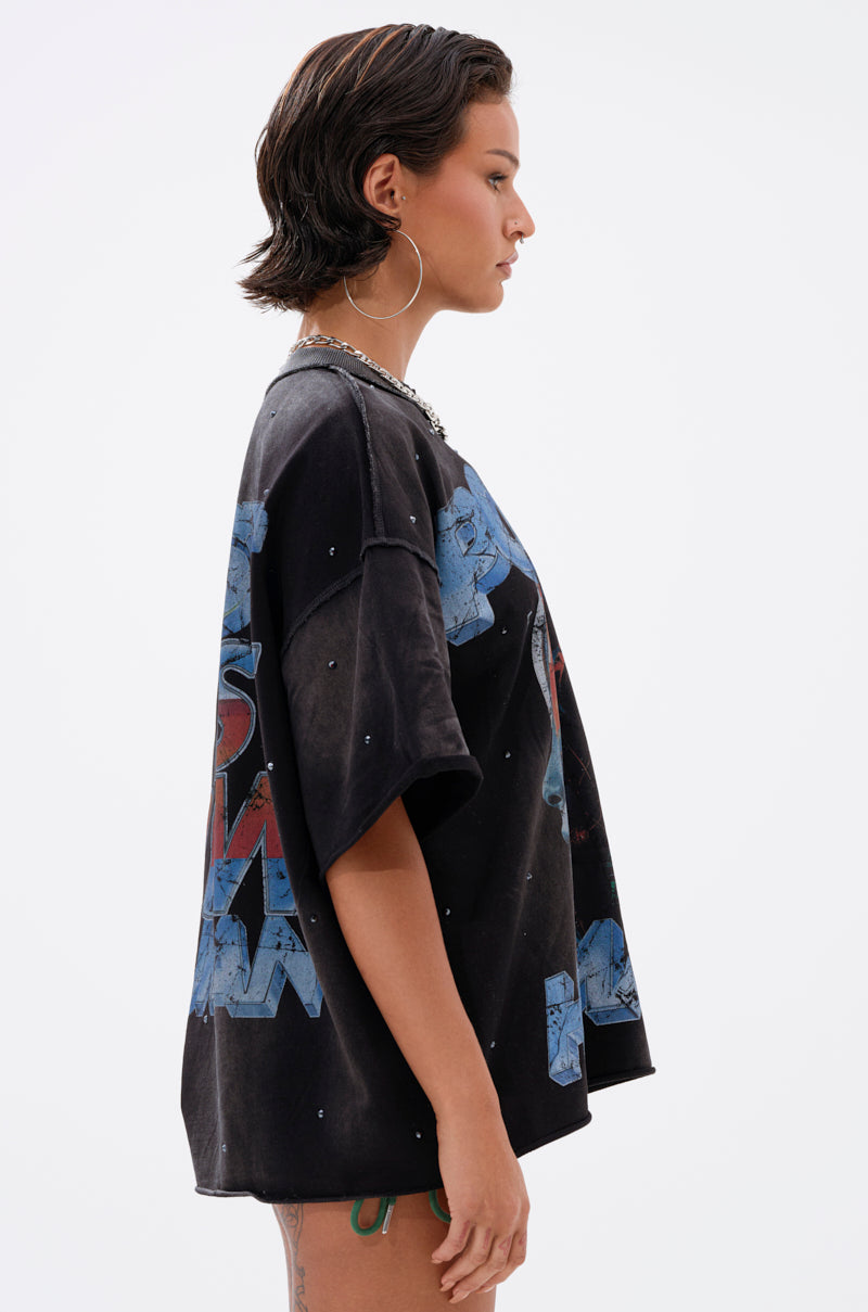 A person with short dark hair and hoop earrings stands in profile against a plain white background, wearing THE FUTURES CALLING FRENCH TERRY GRAPHIC TEE—an oversized shirt with blue and red lettering and light distressing.