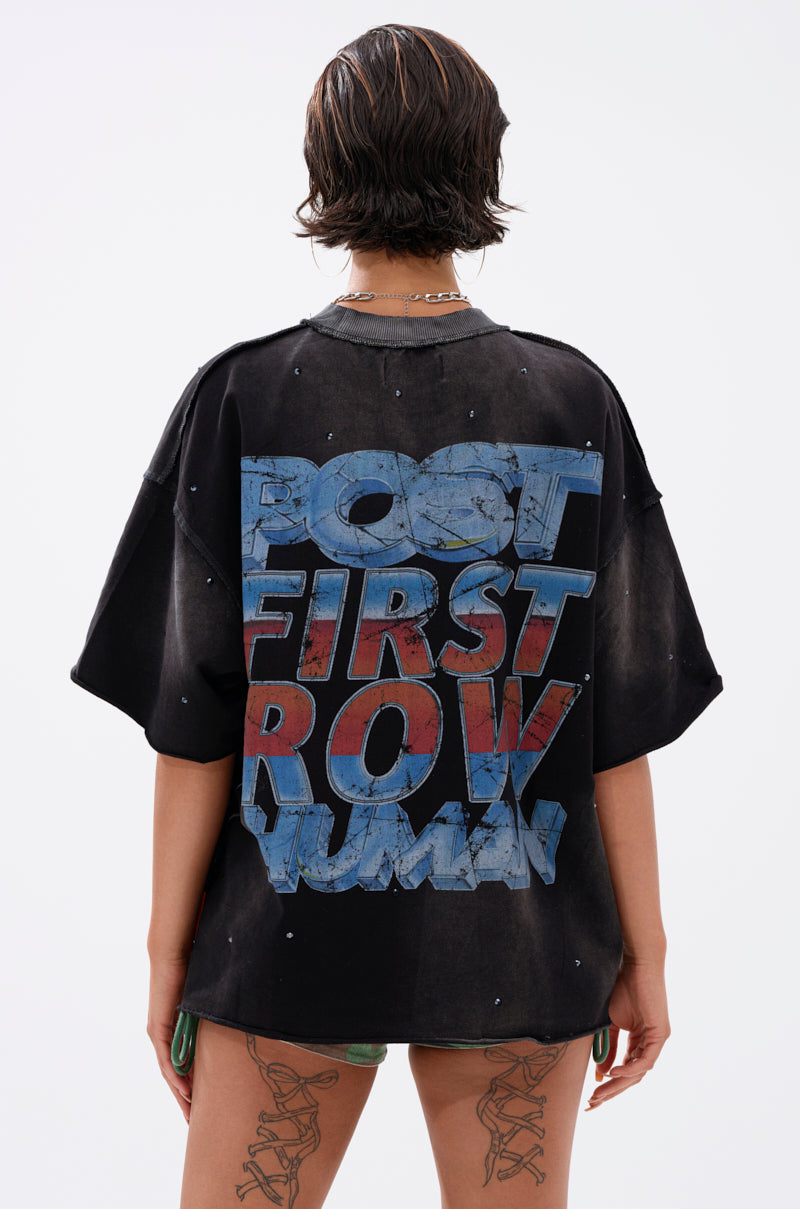 A person with short, wet-look hair stands facing away, wearing THE FUTURES CALLING FRENCH TERRY GRAPHIC TEE—an oversized black shirt with "POST FIRST ROW HUMAN" in bold distressed blue and red on the back, set against a plain white background.