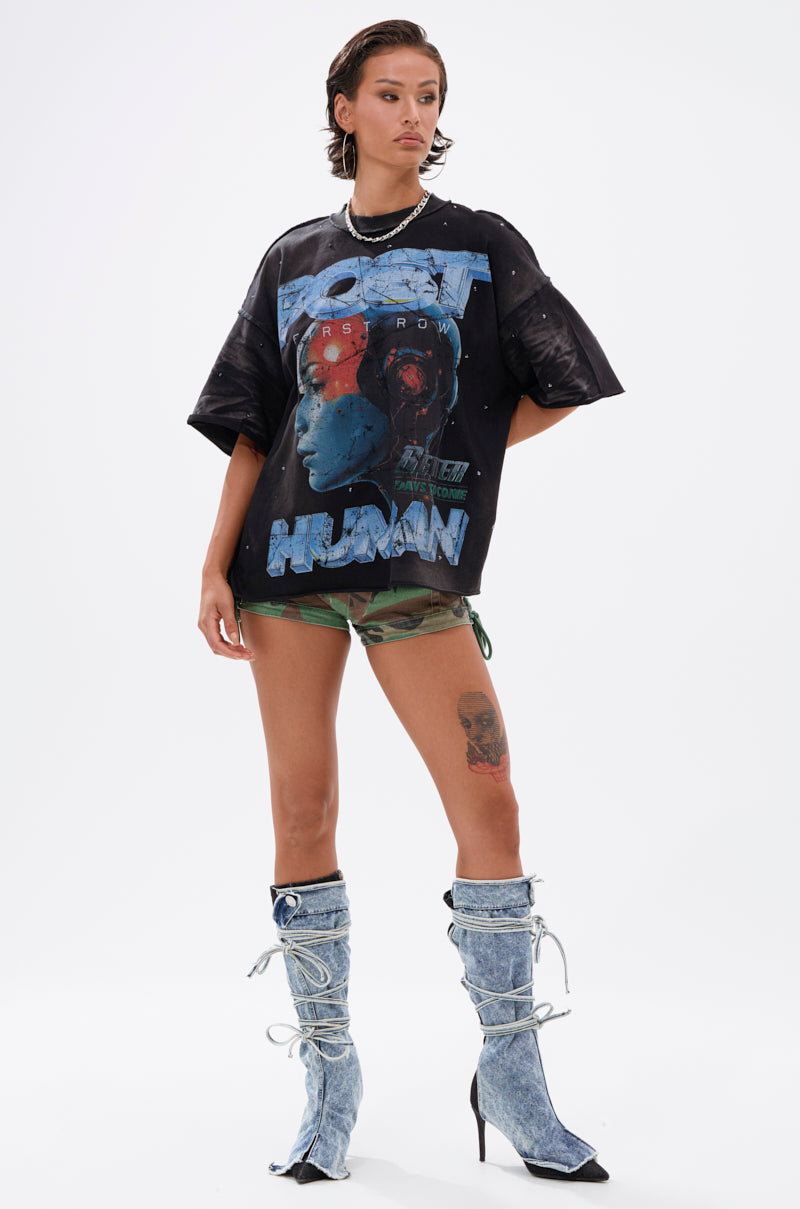 A person with short hair, accessorized with necklaces, wears THE FUTURES CALLING FRENCH TERRY GRAPHIC TEE featuring a futuristic graphic, paired with camouflage shorts and knee-high lace-up denim high-heeled boots against a white background.