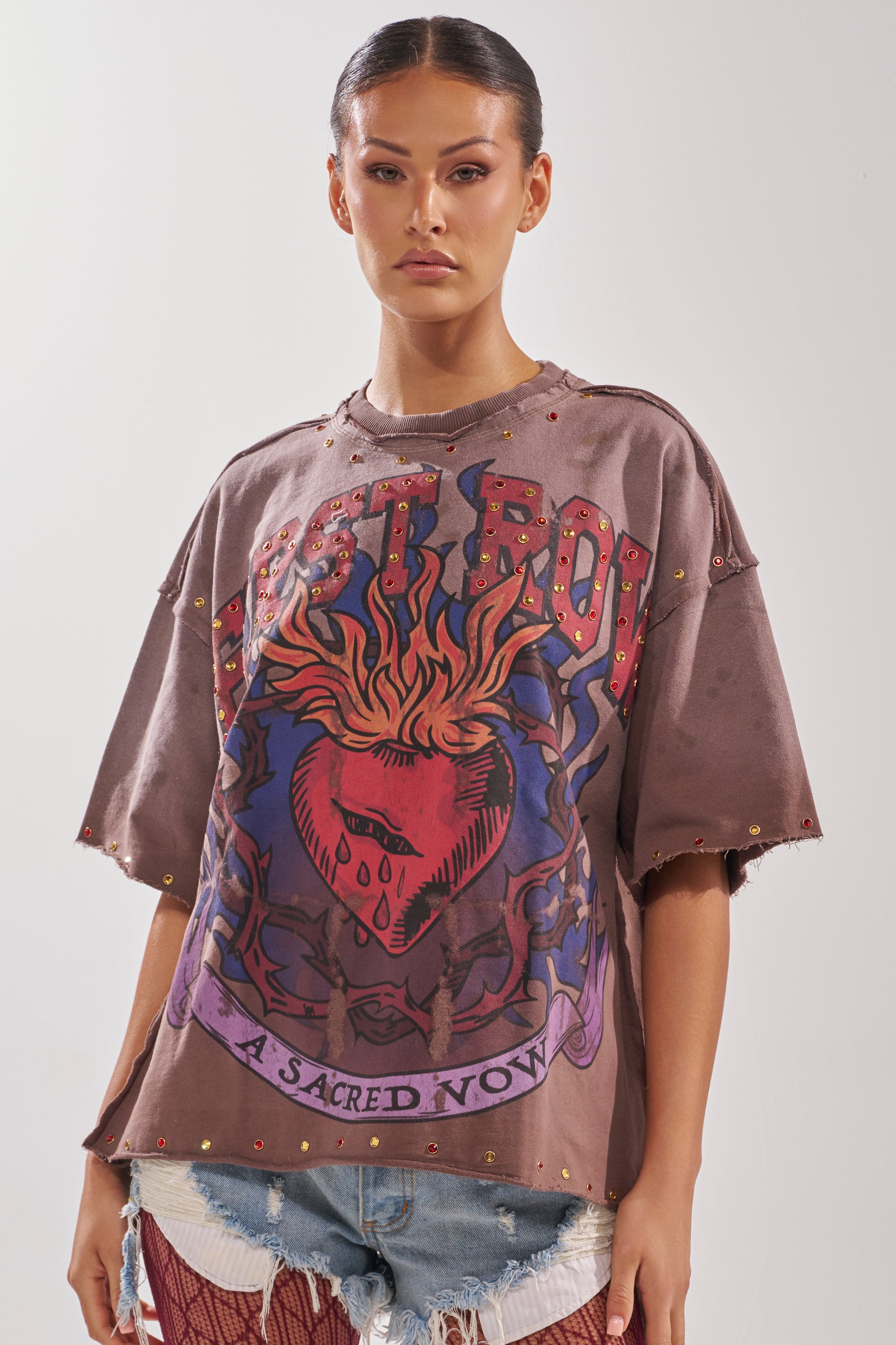 A woman with slicked-back hair wears the SACRED HEARTH RHINESTONE DISTRESSED TEE—an oversized brown graphic t-shirt featuring a bold flaming heart and the words "LOST BOY" and "A SACRED VOW"—paired with distressed denim shorts.