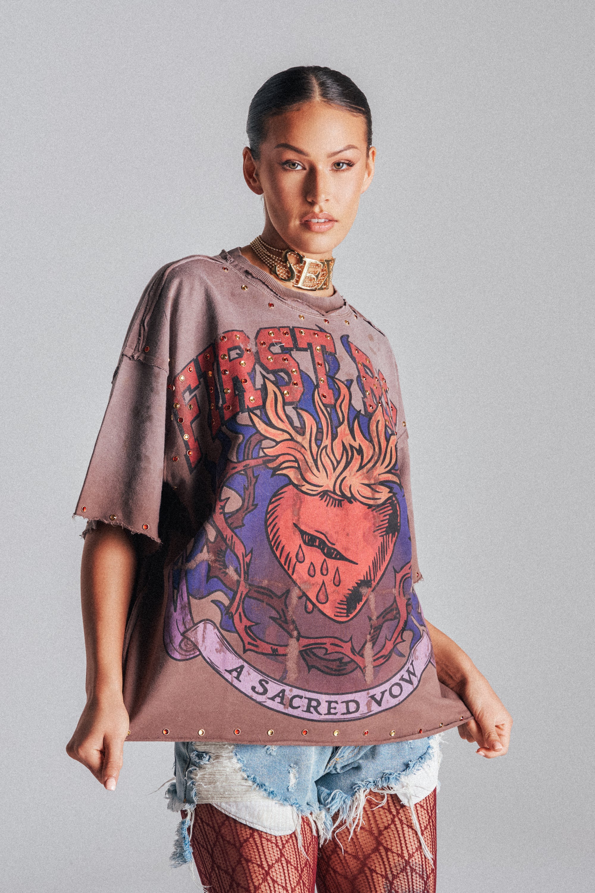 A woman with slicked-back hair poses confidently in the SACRED HEARTH RHINESTONE DISTRESSED TEE, styled with ripped denim shorts, fishnet stockings, and a rhinestone-studded choker against a simple backdrop.