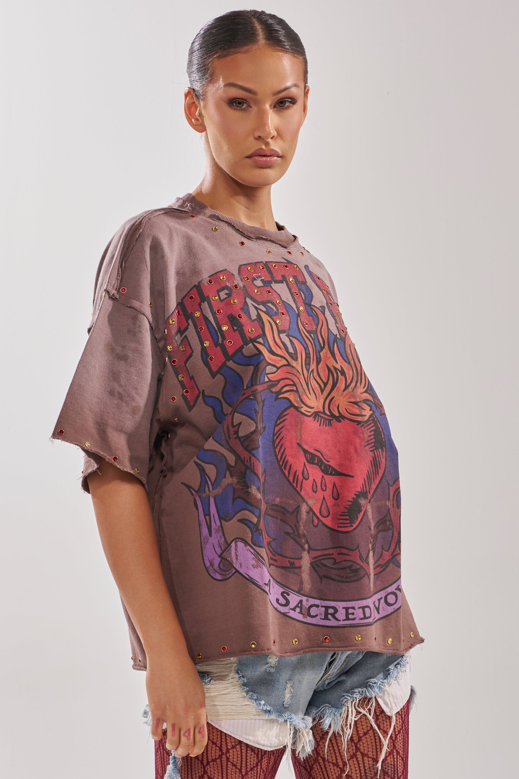 A woman with slicked-back hair channels streetwear in the SACRED HEARTH RHINESTONE DISTRESSED TEE—featuring a flaming heart and "FIRST"—paired with ripped denim shorts and fishnet stockings, standing against a plain background.