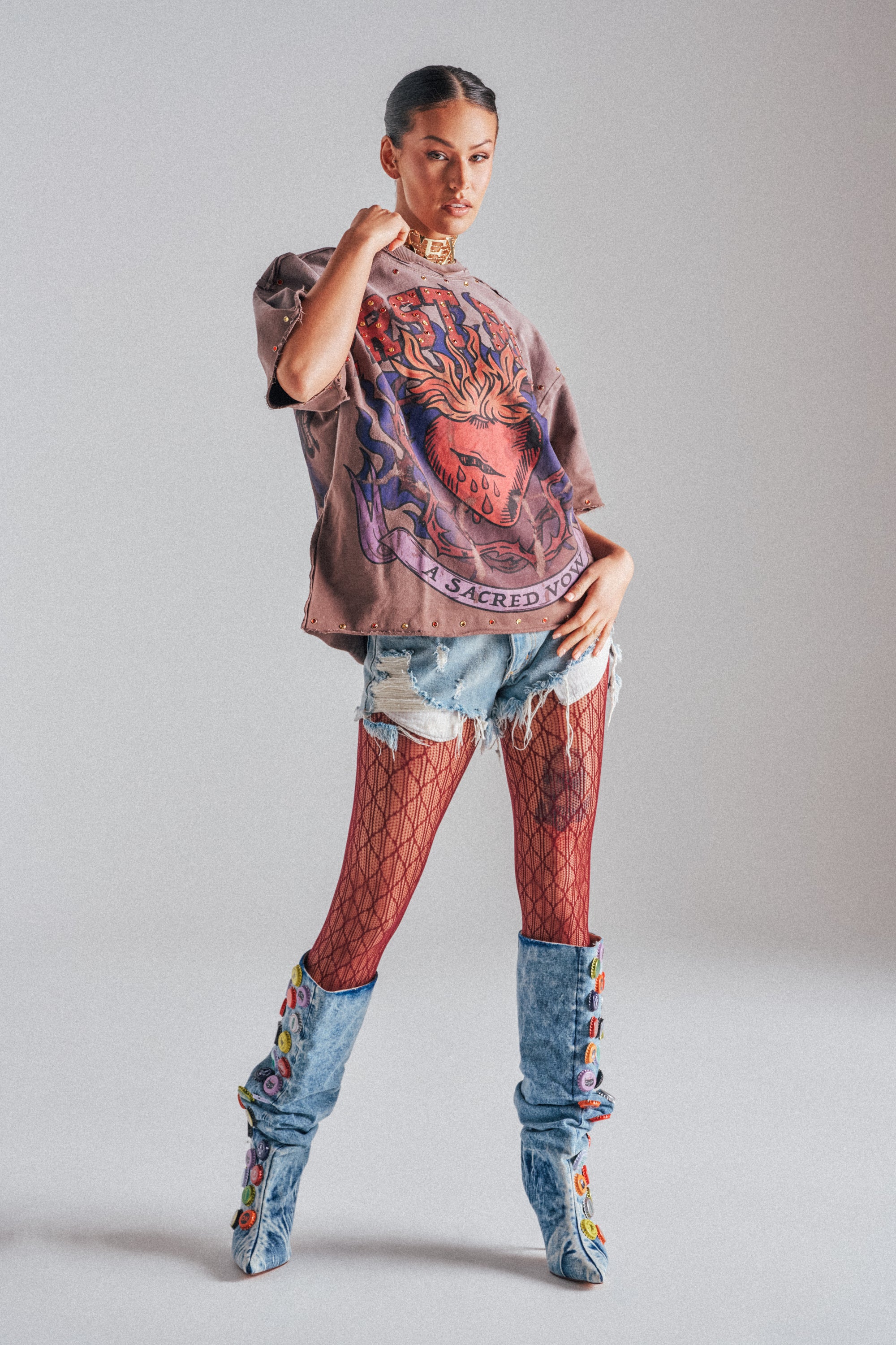 A woman poses confidently in the SACRED HEARTH RHINESTONE DISTRESSED TEE with distressed denim shorts, red fishnet tights, and blue rhinestone-embellished boots, styled against a plain gray background for a bold streetwear look.
