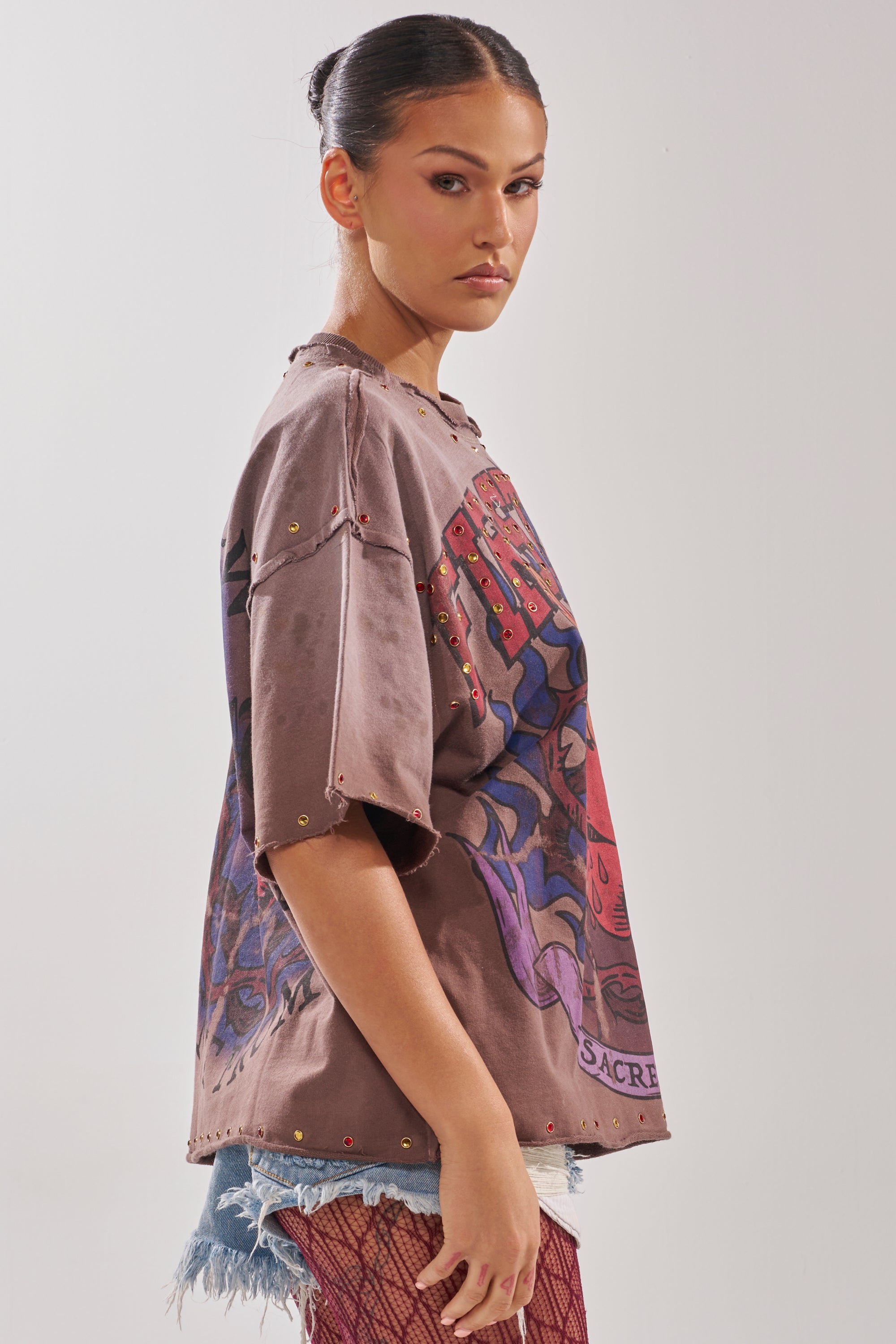 A woman with dark hair in a sleek bun models the SACRED HEARTH RHINESTONE DISTRESSED TEE, styled with distressed denim shorts and red fishnet stockings, against a plain light background.