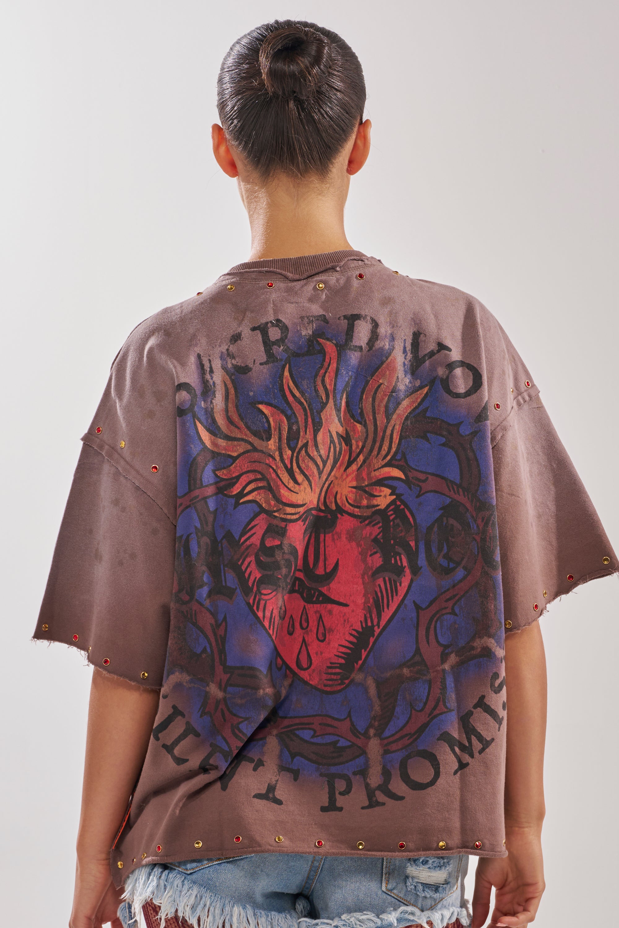 Person with dark hair in a bun, wearing the SACRED HEARTH RHINESTONE DISTRESSED TEE with a large flaming heart and text graphic on the back, and distressed denim shorts, photographed from behind against a plain background.