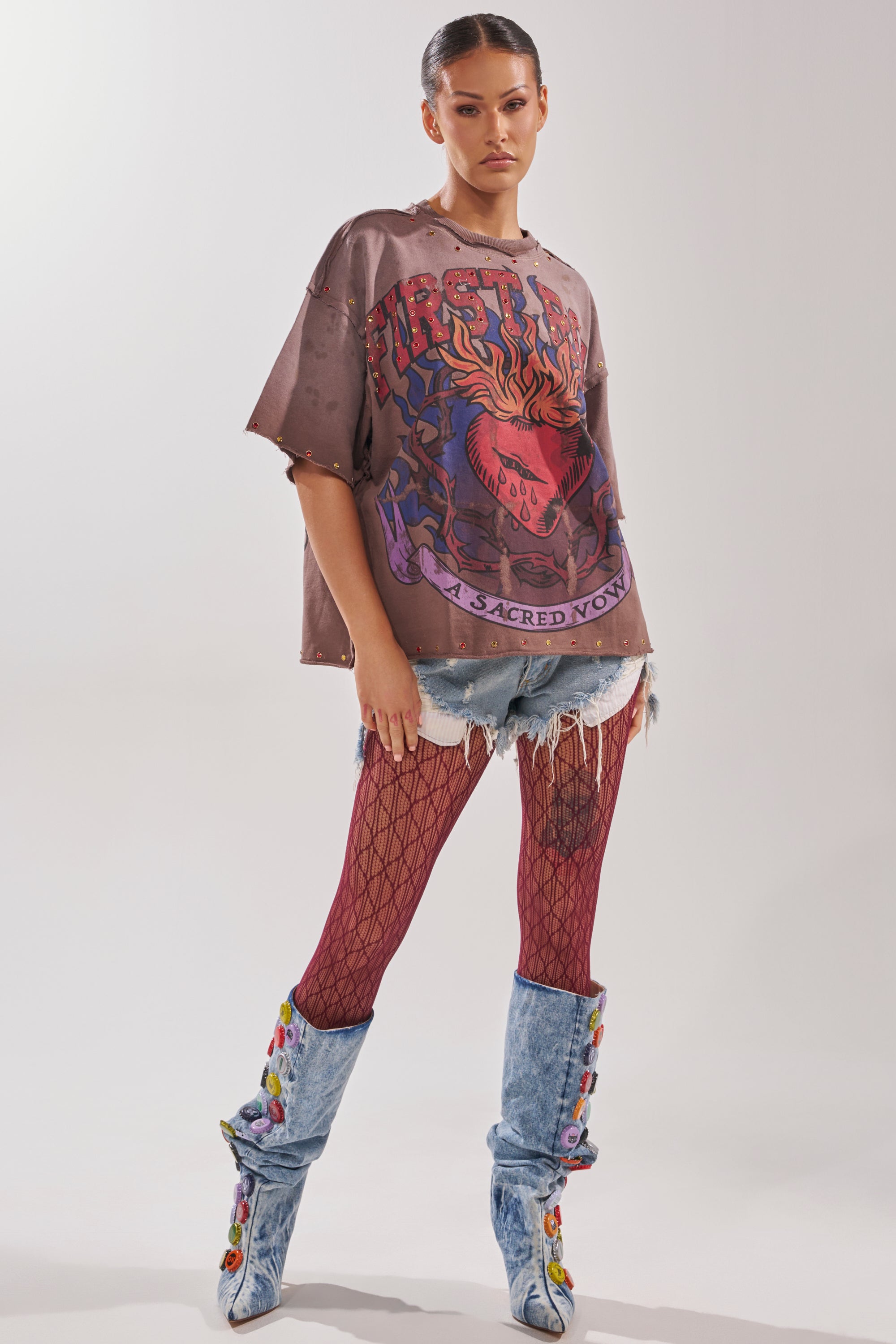 A woman poses confidently in the SACRED HEARTH RHINESTONE DISTRESSED TEE, styled with distressed shorts, red fishnet tights, and light blue rhinestone knee-high boots. Her hair is tied back as she stands against a plain background.