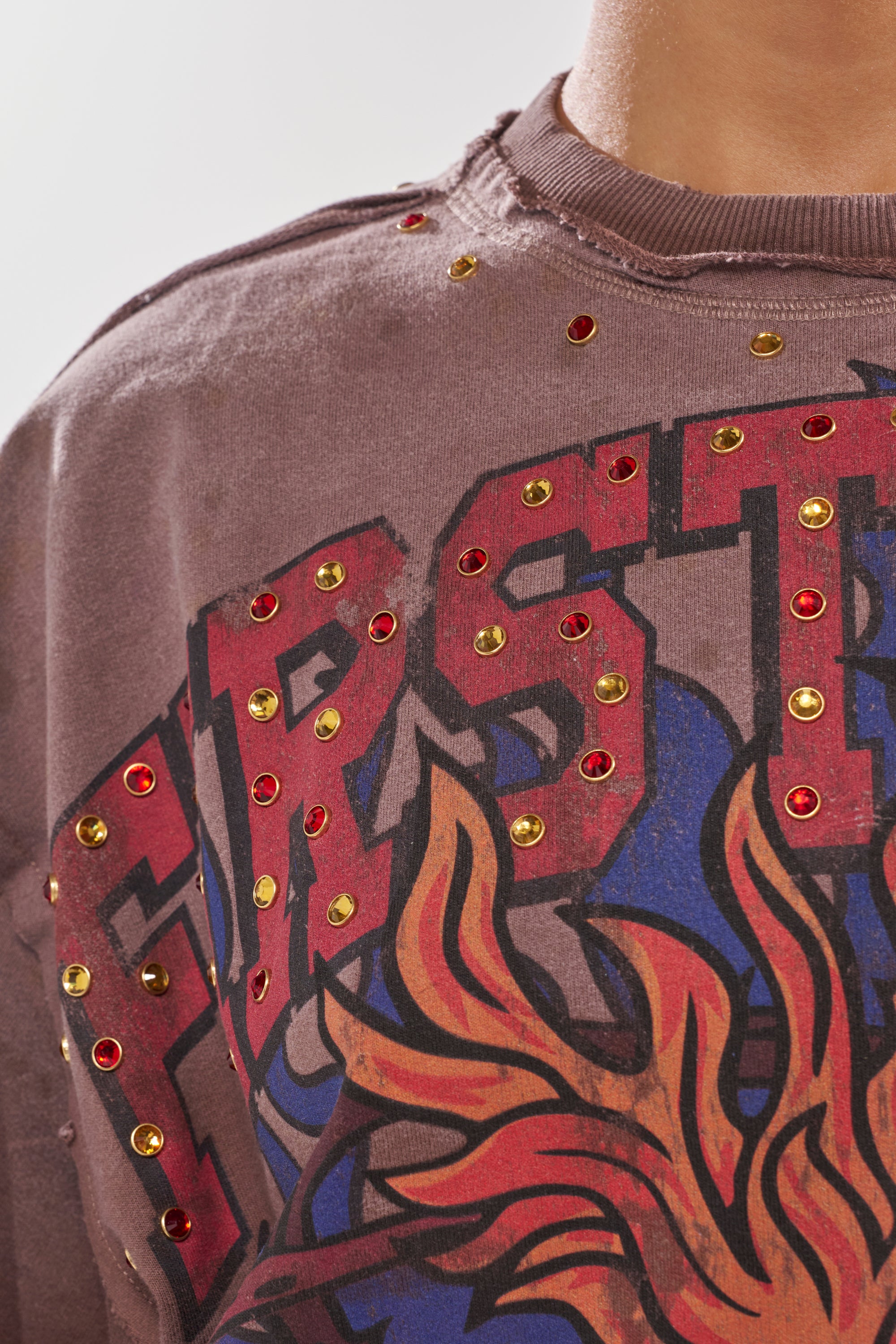A close-up of the SACRED HEARTH RHINESTONE DISTRESSED TEE, a brown graphic t-shirt with large text, colorful flame art, and red and gold streetwear-inspired rhinestone studs accenting the neckline and design.