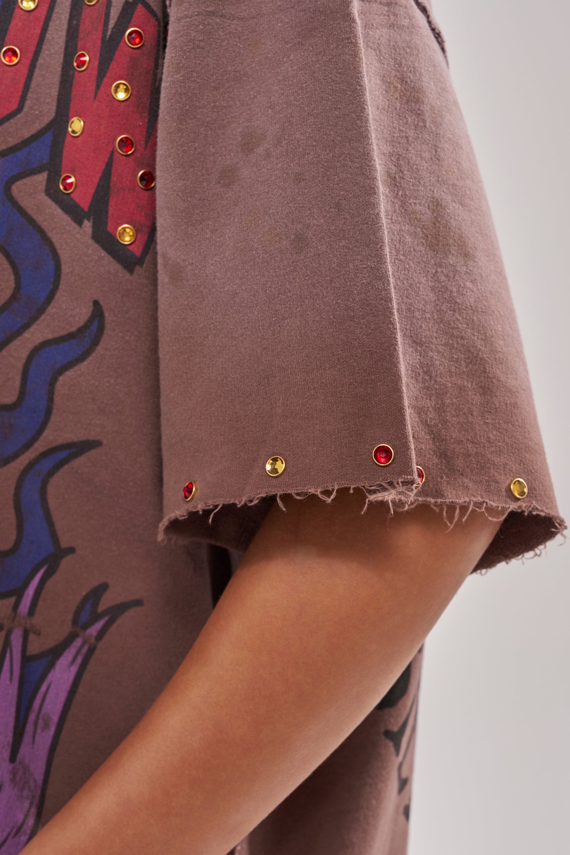 A close-up of a person's arm wearing the SACRED HEARTH RHINESTONE DISTRESSED TEE, a brown streetwear graphic t-shirt with short sleeves, frayed hem, rhinestone detailing, and a vibrant red and blue design.