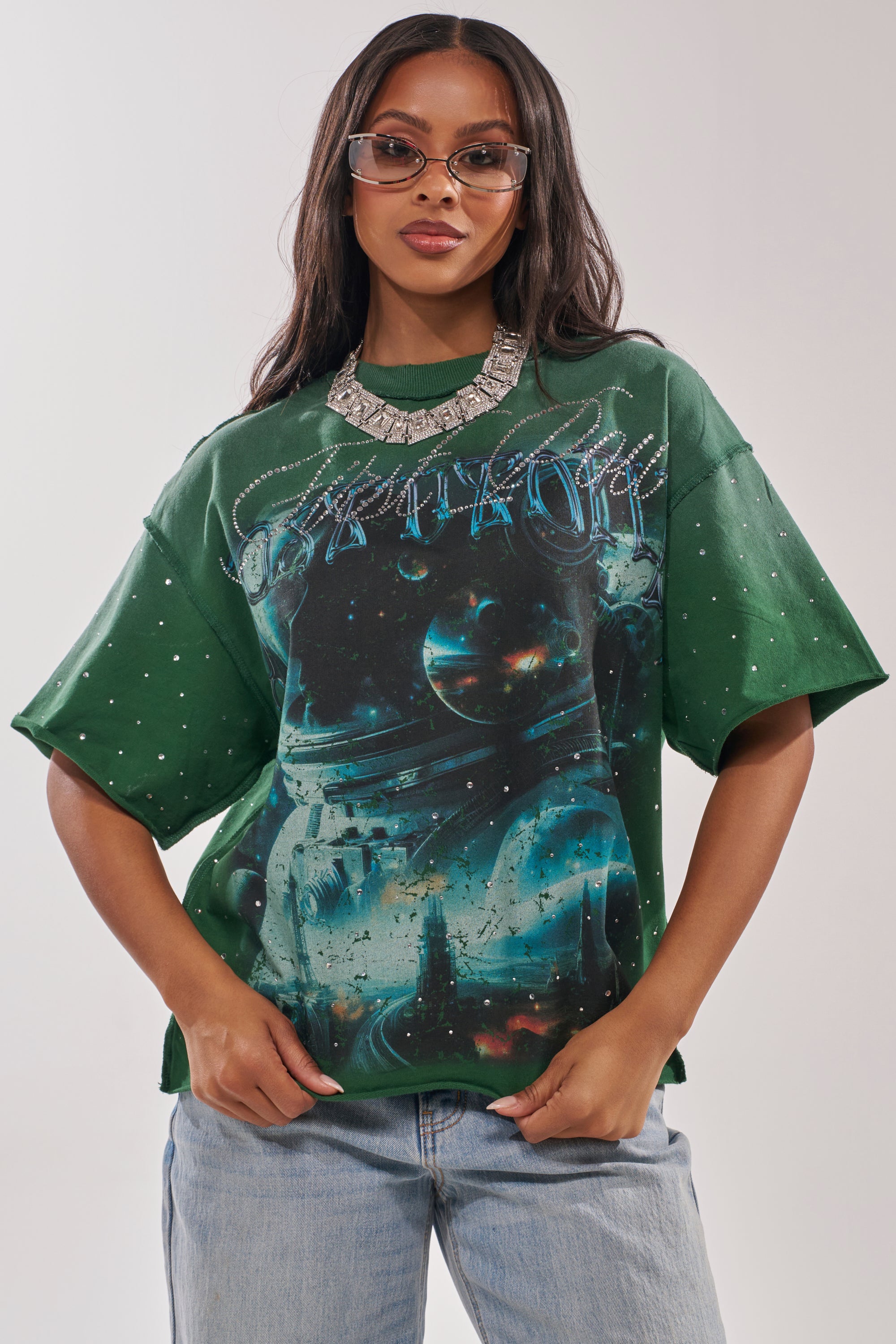 A woman in streetwear style stands confidently, wearing oversized sunglasses, a chunky silver necklace, light blue jeans, and the FIRST ROW INTERSTELLAR RHINESTONE FRENCH TERRY TEE with a cosmic print and rhinestone accents. 0