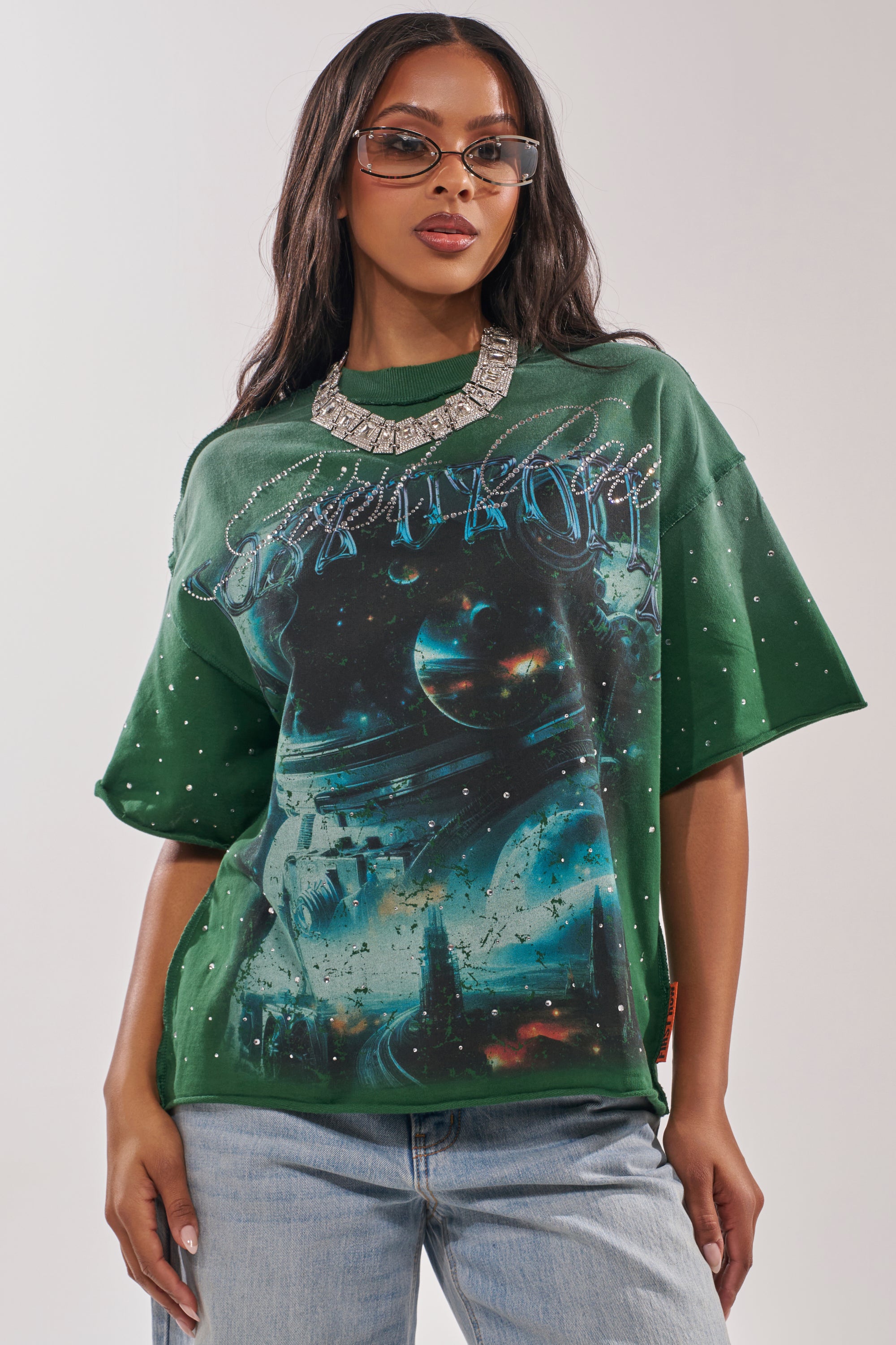 Wearing sunglasses and a chunky silver necklace, a woman poses against a plain background in the FIRST ROW INTERSTELLAR RHINESTONE FRENCH TERRY TEE, an oversized space-themed graphic top with rhinestones, paired with trendy light blue jeans. 1