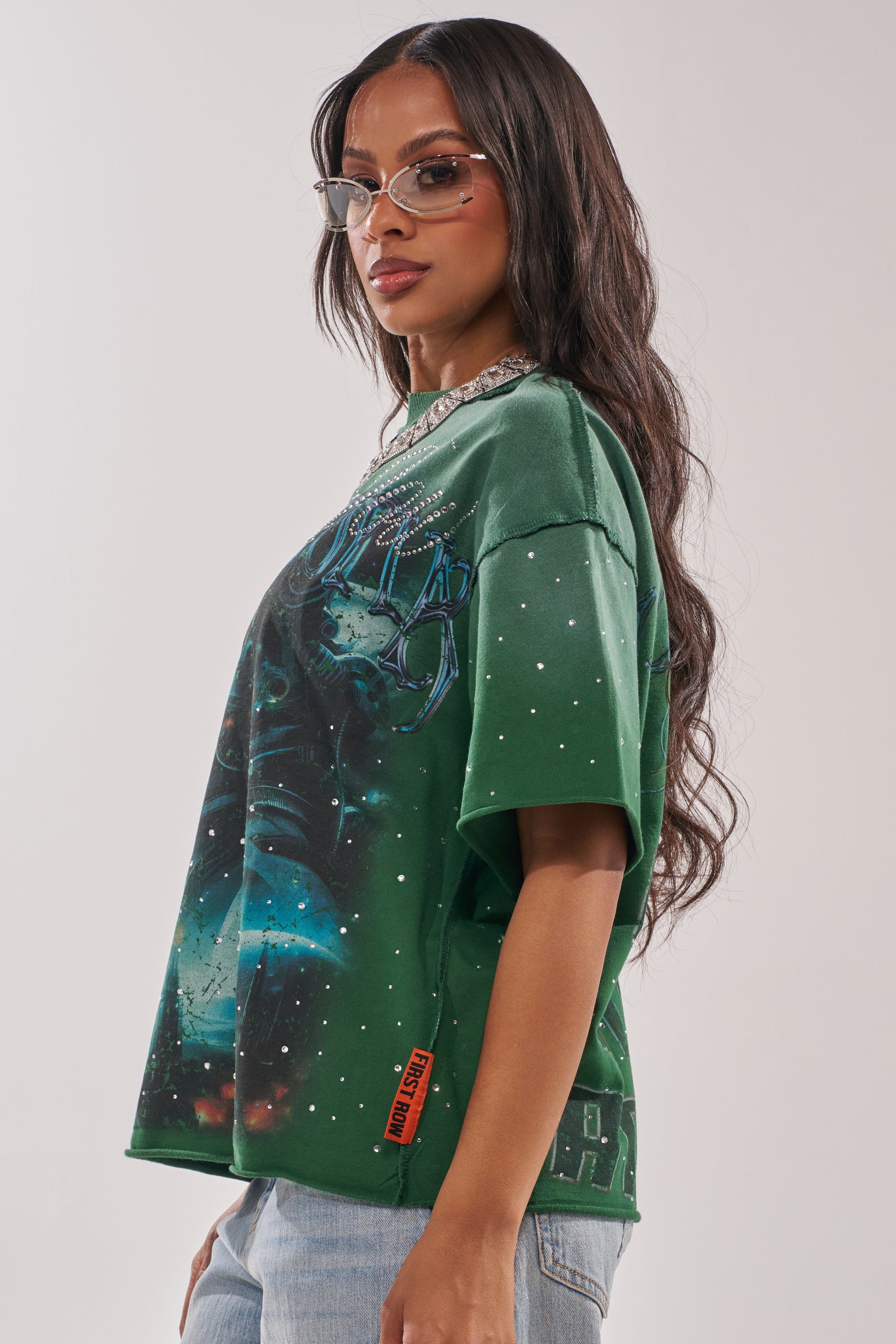 A woman with long, wavy dark hair and stylish glasses poses sideways in the FIRST ROW INTERSTELLAR RHINESTONE FRENCH TERRY TEE, featuring a galaxy design with rhinestone embellishments, paired with a chunky necklace and light blue jeans. 2