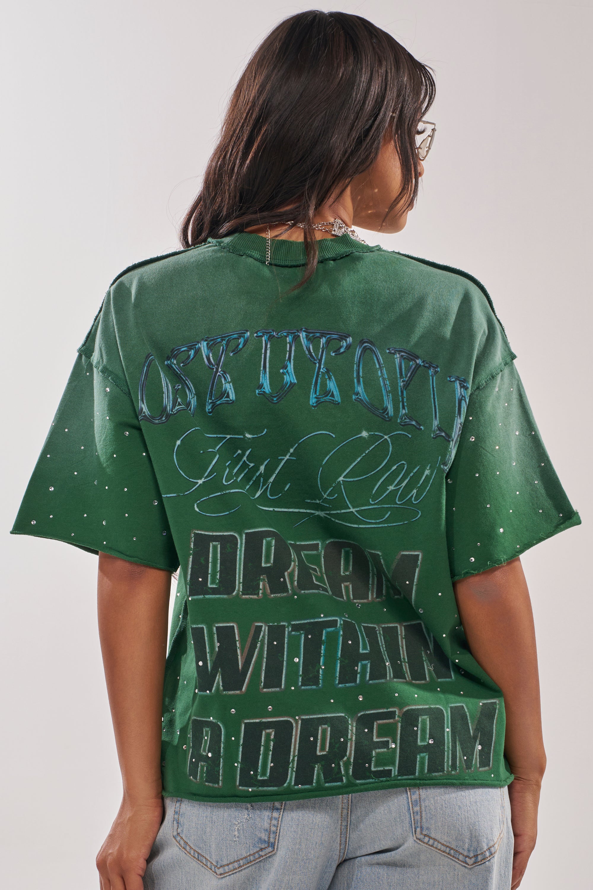 A woman with long dark hair and glasses is seen from the back wearing the FIRST ROW INTERSTELLAR RHINESTONE FRENCH TERRY TEE in green, featuring “DREAM WITHIN A DREAM” text, styled with light denim jeans for a streetwear look. 3