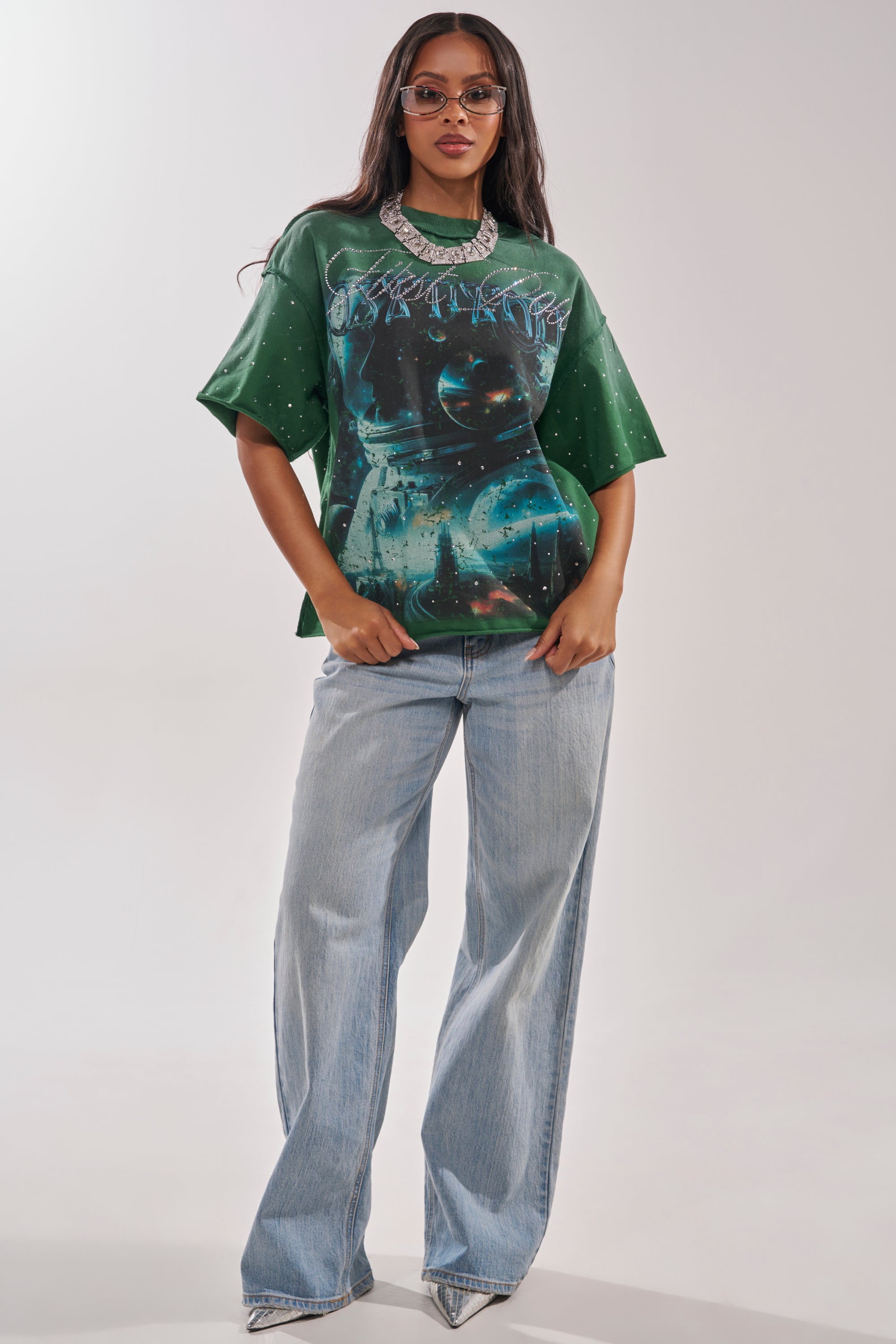 A woman stands confidently against a plain light background, wearing sunglasses, a chunky necklace, light blue wide-leg jeans, and the FIRST ROW INTERSTELLAR RHINESTONE FRENCH TERRY TEE. 4