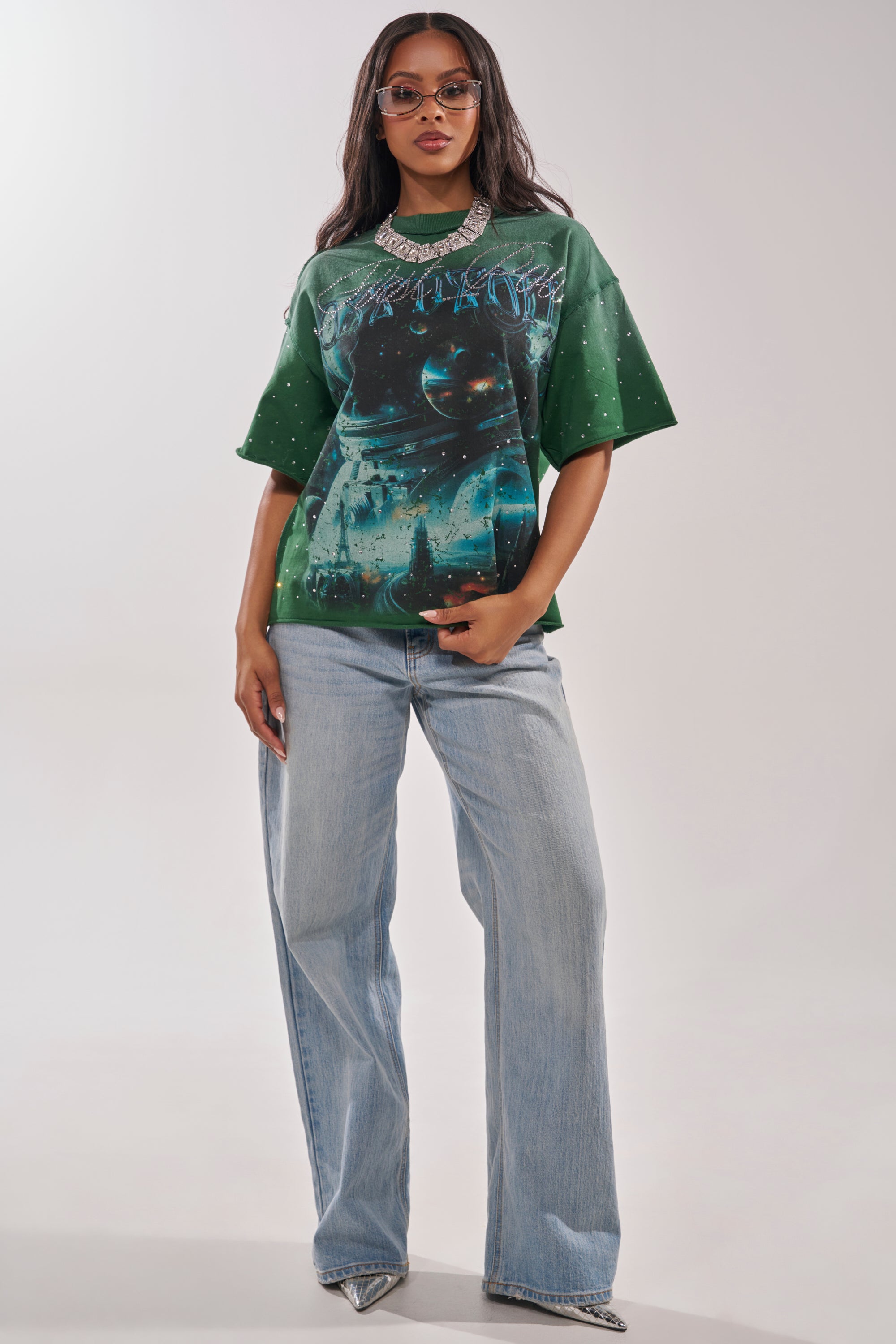 A woman styles the FIRST ROW INTERSTELLAR RHINESTONE FRENCH TERRY TEE—an oversized space-print graphic tee with rhinestones—with light wash wide-leg jeans, a crystal necklace, glasses, and silver metallic heels for a bold streetwear vibe. 5