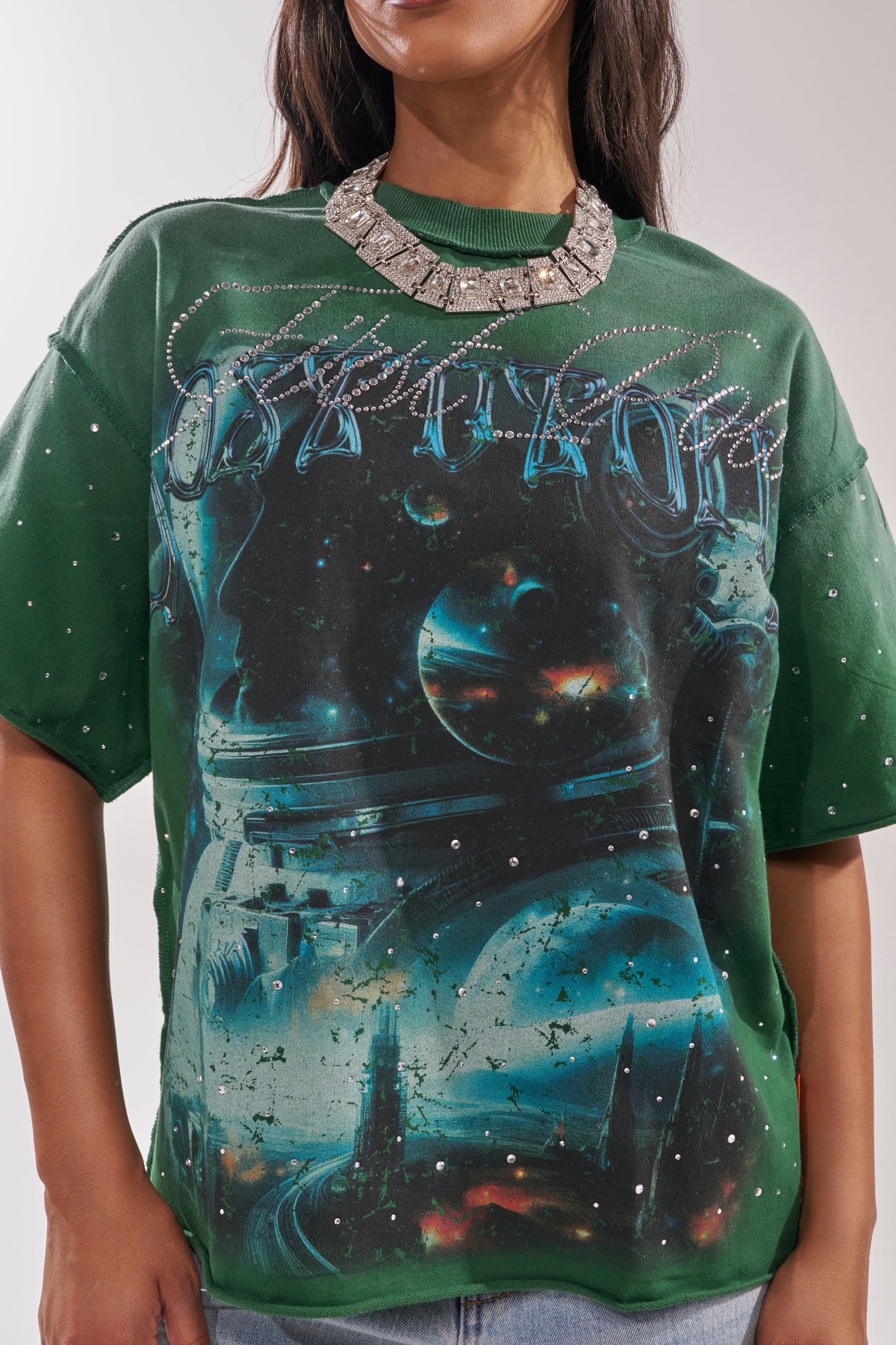 A woman wears the FIRST ROW INTERSTELLAR RHINESTONE FRENCH TERRY TEE, an oversized green graphic shirt with a vibrant outer space design, paired with a bold silver necklace for standout streetwear style. 7