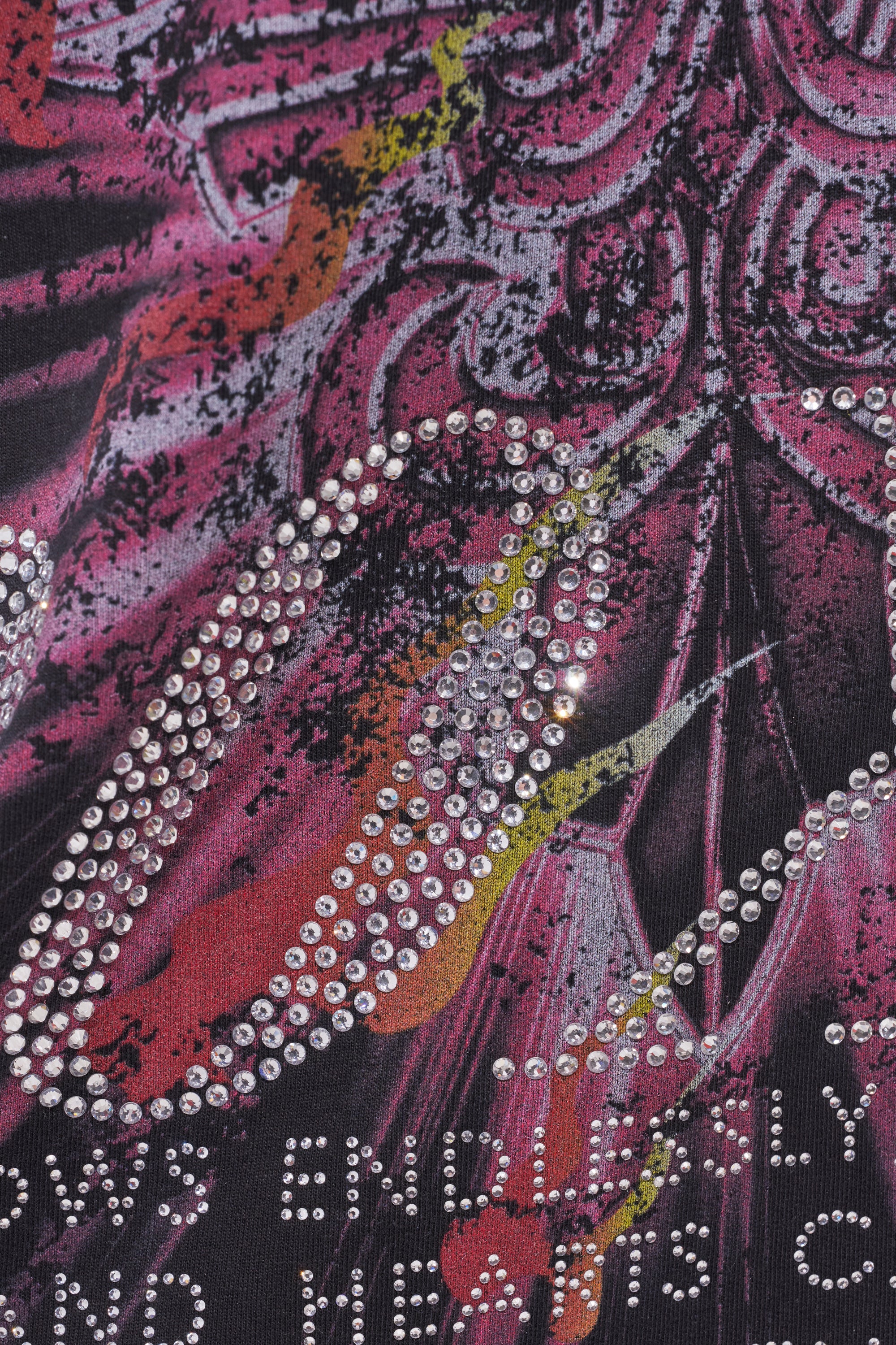 Close-up of the 20XX RHINESTONE CANVAS T-SHIRT, featuring a black cotton base with a colorful abstract pattern in red, purple, yellow, and black, accented by rows of reflective rhinestones and partially visible white text at the hem. 8