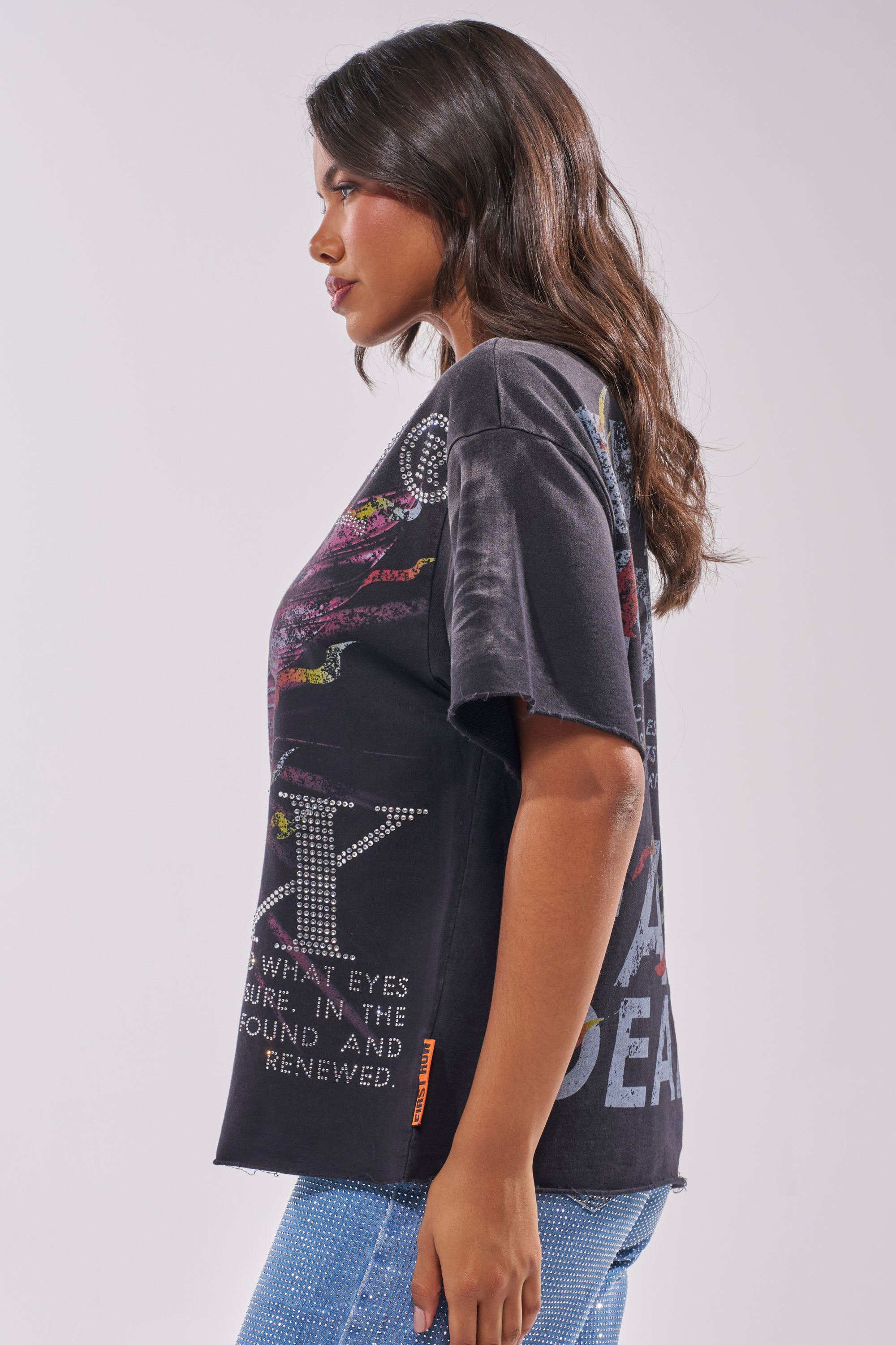 A woman with wavy brown hair stands in profile against a plain light background, wearing the 20XX RHINESTONE CANVAS T-SHIRT—a loose black graphic tee with rhinestone accents—paired with light blue jeans. 2