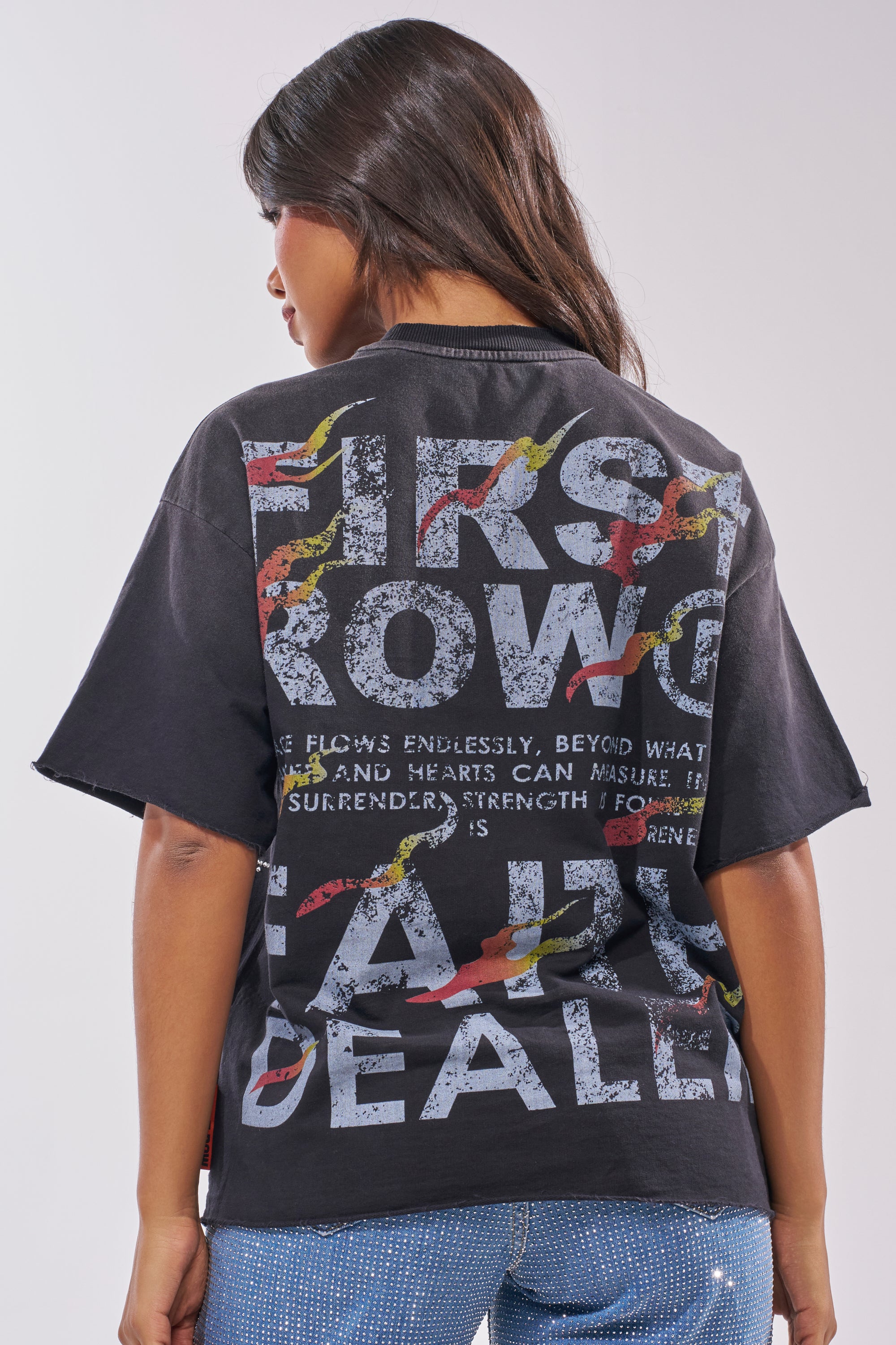 A woman with long dark hair faces away from the camera, wearing the 20XX RHINESTONE CANVAS T-SHIRT in washed black cotton, featuring bold white text and colorful graphics on the back, set against a plain light gray background. 3