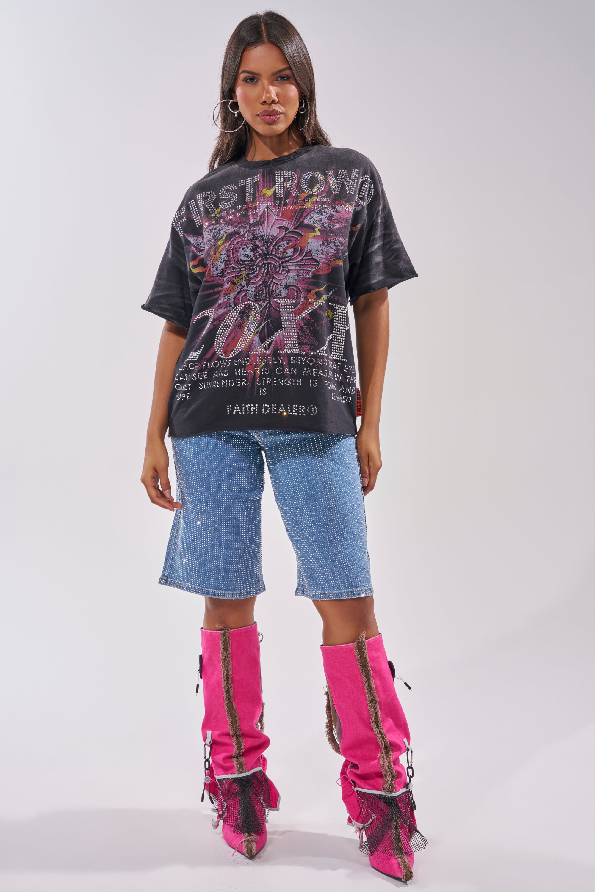 A woman with long dark hair and hoop earrings wears the 20XX RHINESTONE CANVAS T-SHIRT in washed black, blue knee-length denim shorts, and bright pink knee-high boots with black mesh details against a light gray background. 7