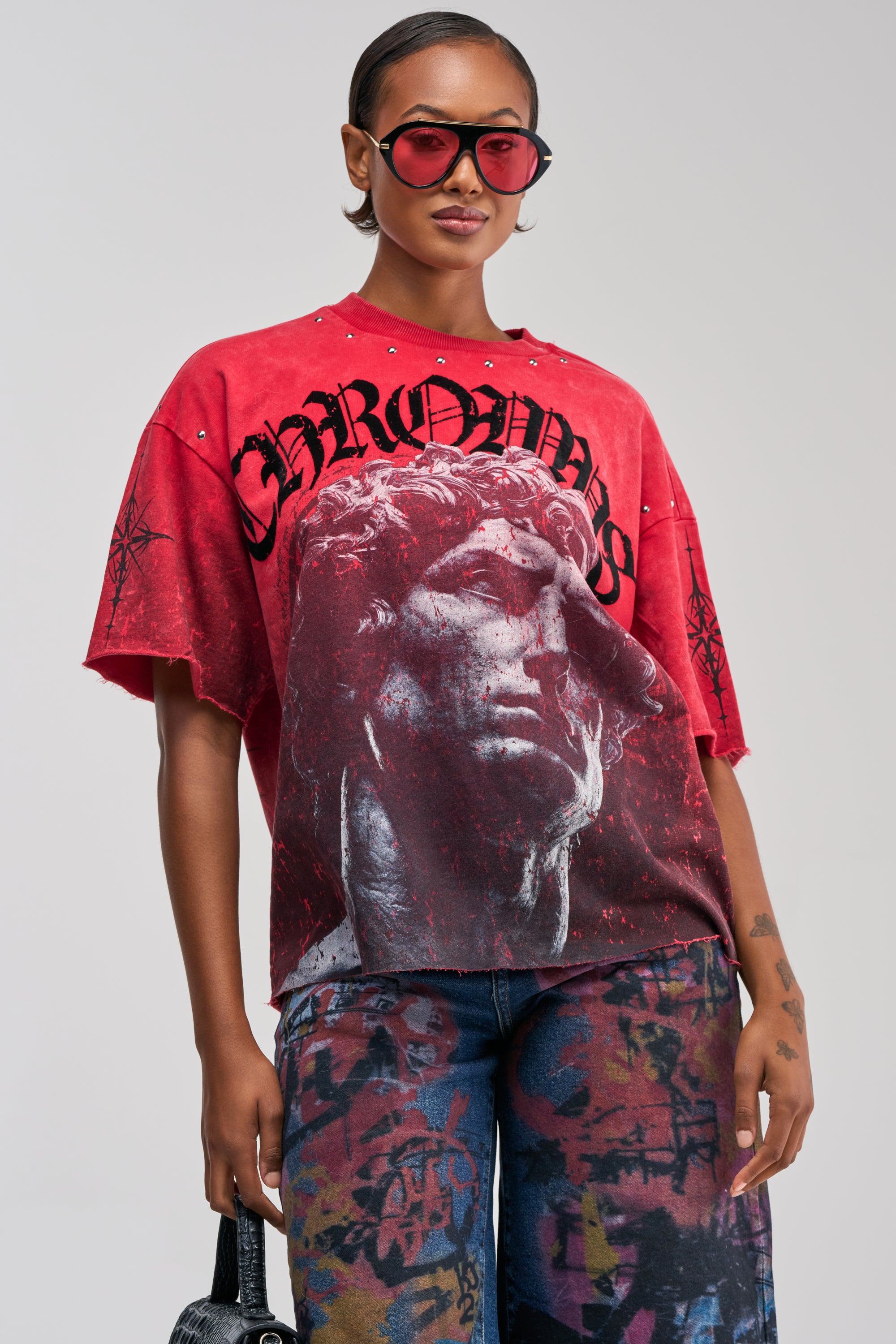 Wearing red sunglasses and the FIRST ROW ONLY HUMAN GRAPHIC TEE—an oversized, relaxed fit shirt with a bold graphic and statement text—a person stands confidently against a plain background, completing their streetwear look with vibrant graffiti-style pants.