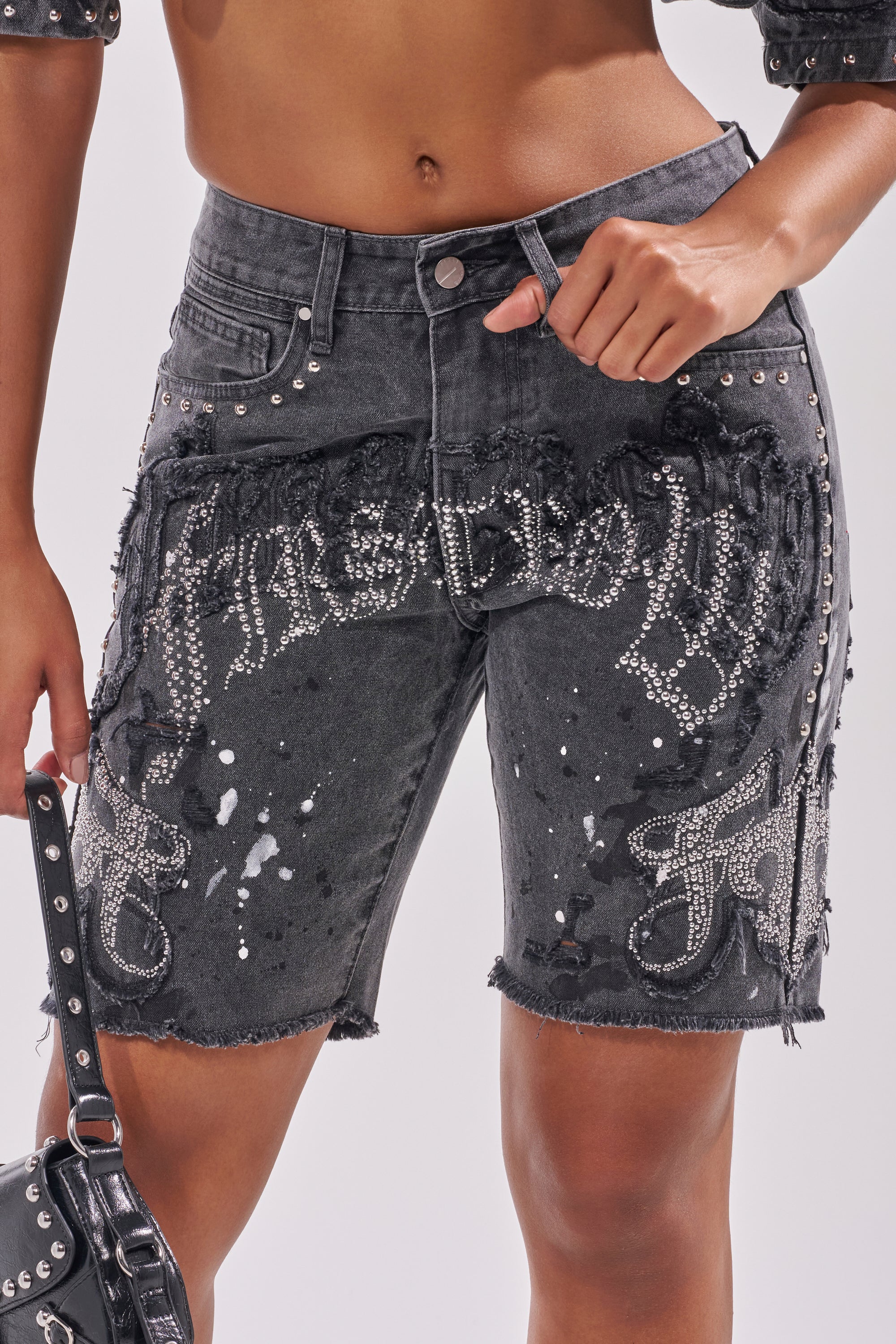 Wearing the NOT LIKE THE REST EMBELLISHED DENIM SHORT—high-waisted black denim with frayed edges, silver studs, rhinestones, and white splatters—paired with a studded black bag for a bold concert look.
