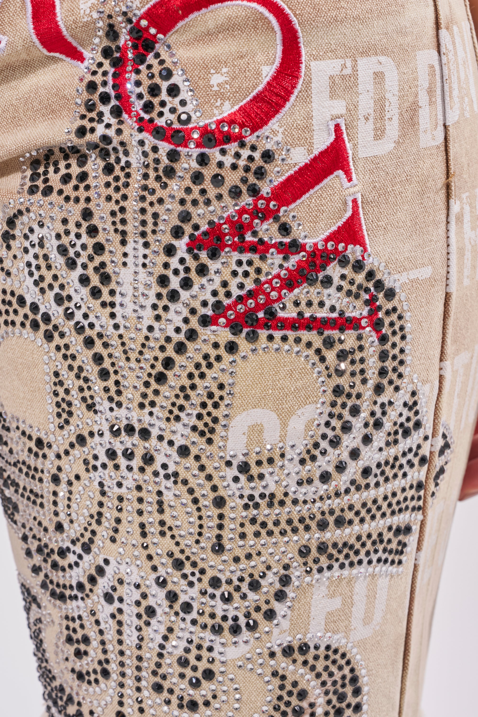 Close-up of the LIGHT SPEAKS IN SILENCE DENIM SHORT in beige, featuring ornate black rhinestone designs, white printed text, and bold red embroidered letters for a textured, decorative streetwear look. 11