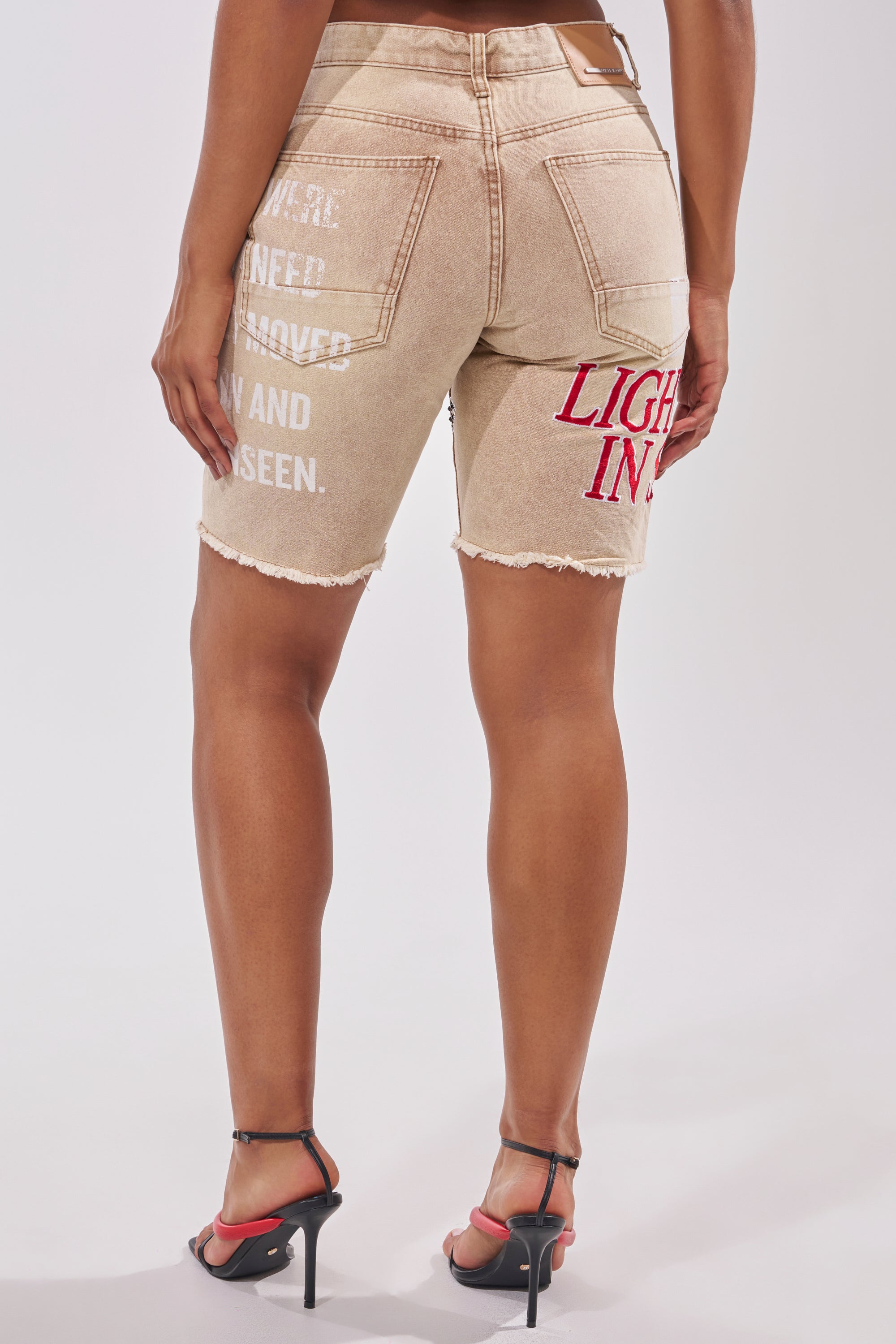 A person wears the LIGHT SPEAKS IN SILENCE DENIM SHORT, featuring tan distressed denim, printed phrases like "NEED APPROVED AND SEEN" (white) and "LIGHT IN" (red) on the back right, styled with red heels for a standout streetwear look. 7