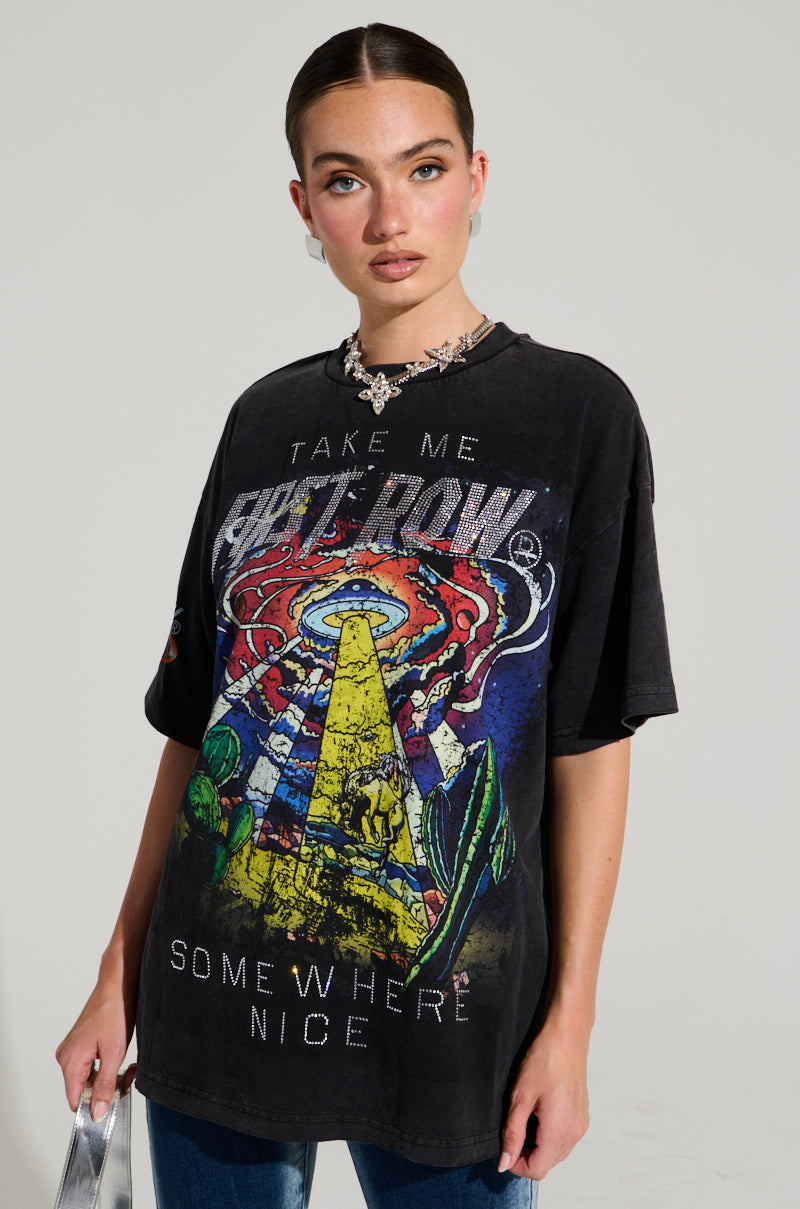 A model with tied-back hair and hoop earrings showcases the TAKE ME AWAY TSHIRT, a black oversized top featuring cowboy-inspired graphic designs. The shirt displays a lighthouse sending a beam to an alien spaceship amidst cactus, flames, and rhinestone-adorned lettering. They complement the look with jeans and hold a silver bag.