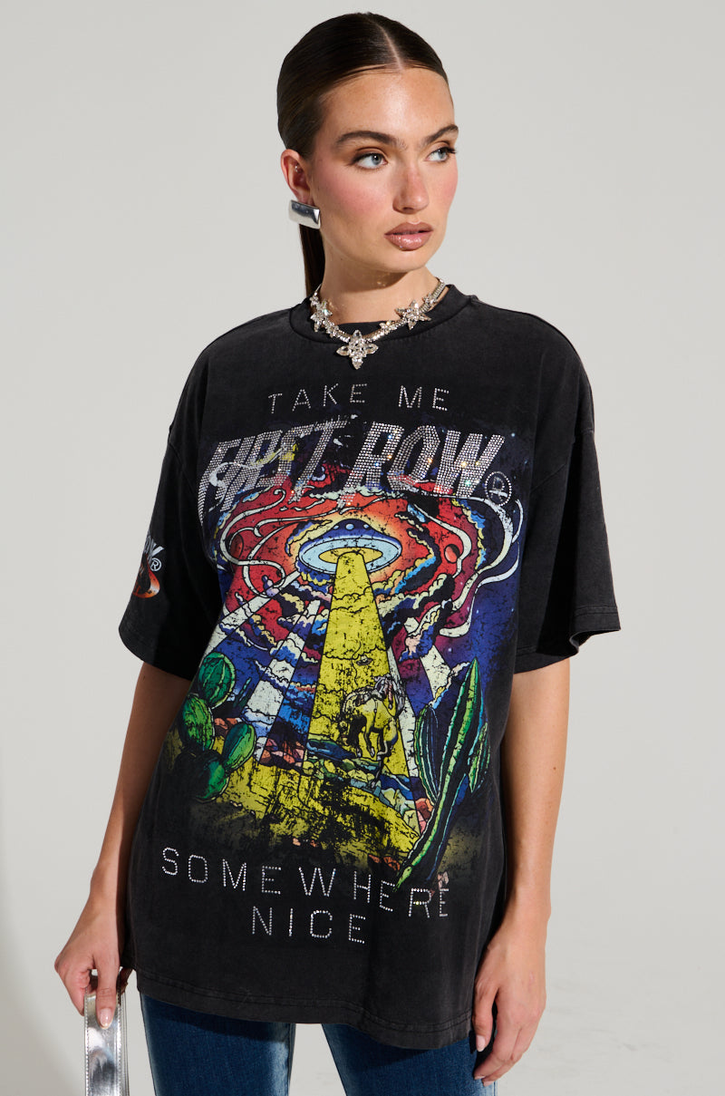 A woman wears the FIRST ROW MIDNIGHT RHINESTONE EMBELLISHED T-SHIRT featuring vibrant UFO and cowboy graphics that illuminate a desert. The rhinestone-adorned "Take Me Somewhere Nice" lettering sparkles as she pairs it with a chunky necklace and silver handbag.